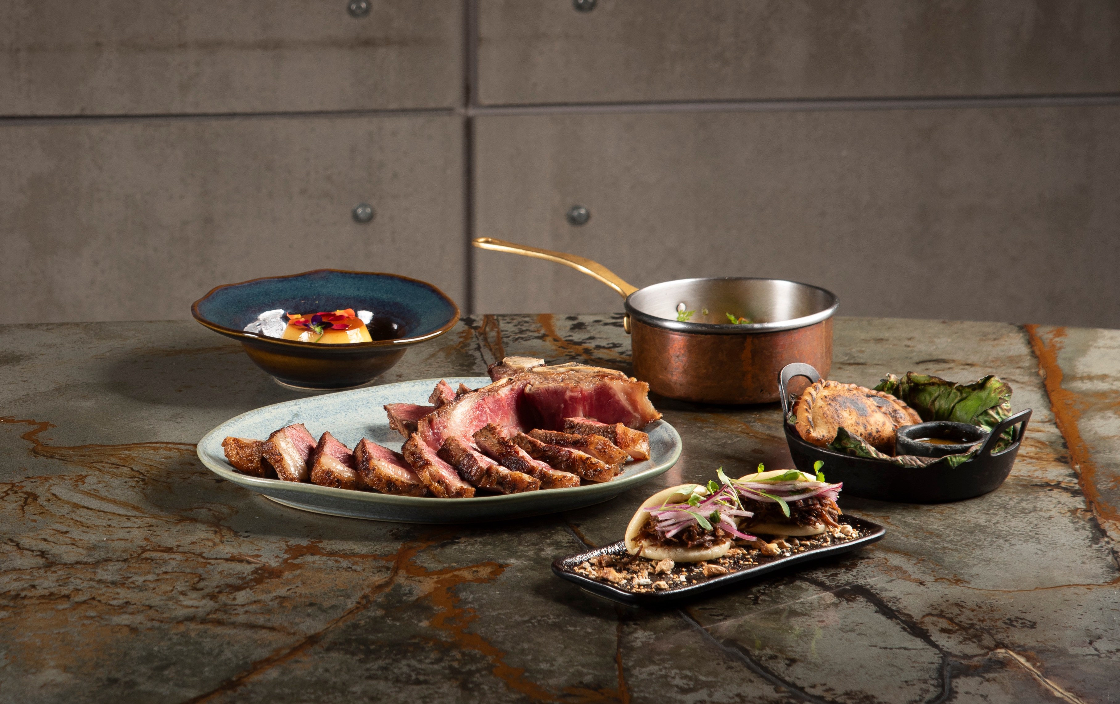 Mesa com pratos variados: um bife fatiado ao centro, um doce em tigela azul, um molho em panela de cobre, e dois p&atilde;es recheados com carne e cebola roxa