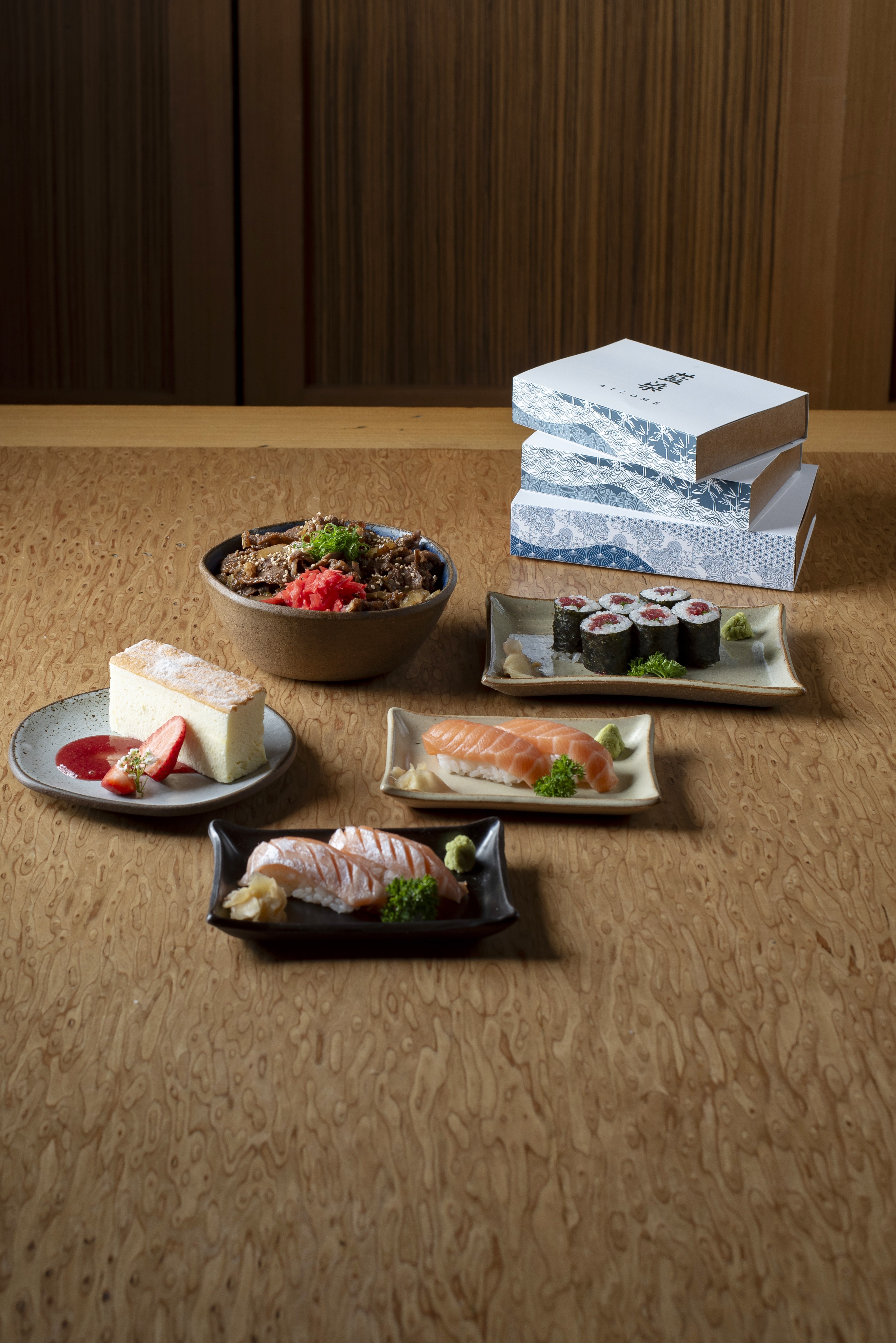 Uma variedade de pratos japoneses dispostos sobre uma mesa de madeira clara. H&aacute; um bowl de gyudon, um peda&ccedil;o de cheesecake com calda de morango, um prato com seis hossomakis, um prato com dois niguiris de salm&atilde;o e outro com dois sushis de salm&atilde;o ma&ccedil;aricado. Ao fundo, tr&ecirc;s caixas brancas empilhadas com desenhos azuis de ondas e montanhas