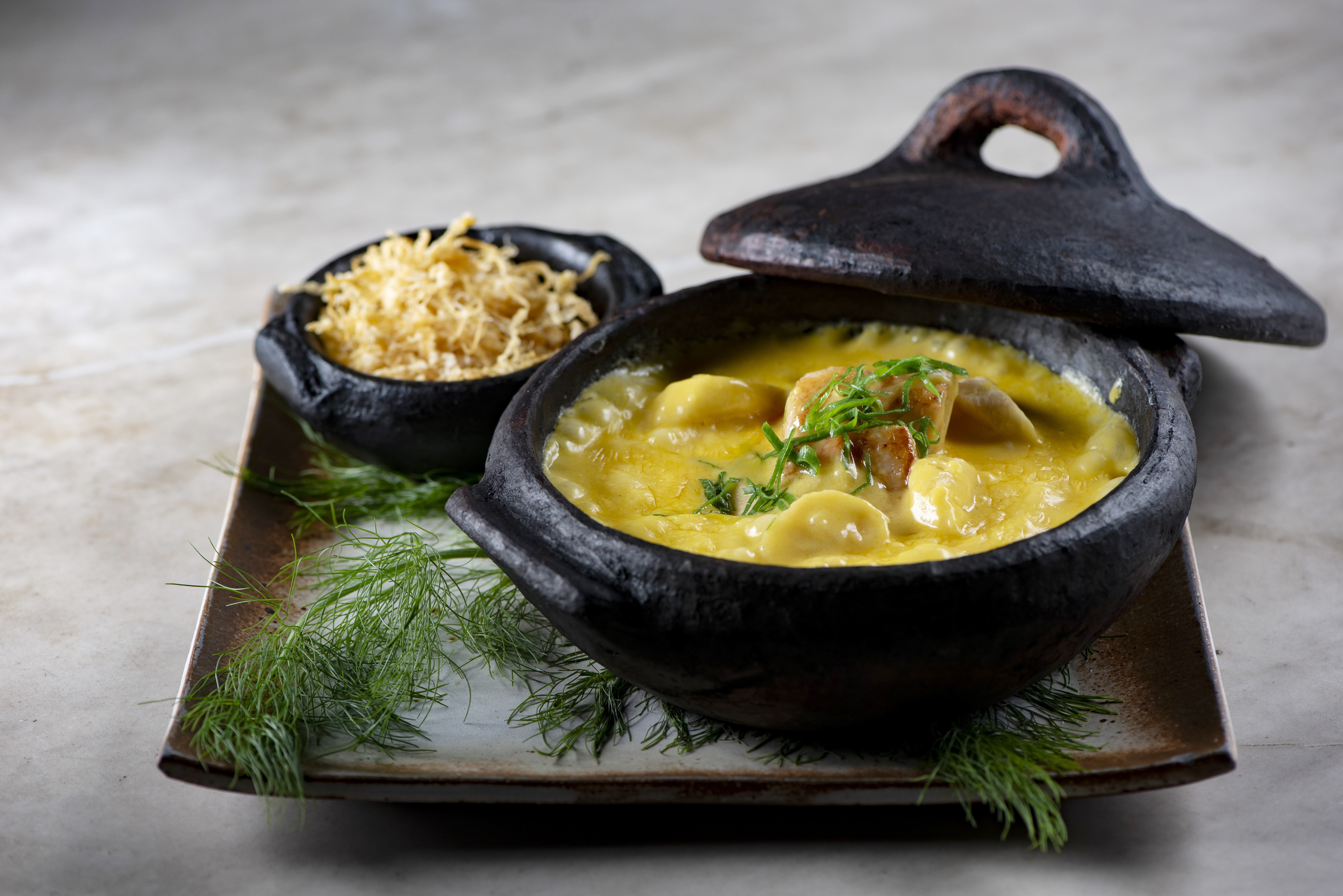 Moqueca de peixe em panela de barro preta com tampa, acompanhada de farofa crocante em tigela menor, sobre bandeja r&uacute;stica com ramos de endro