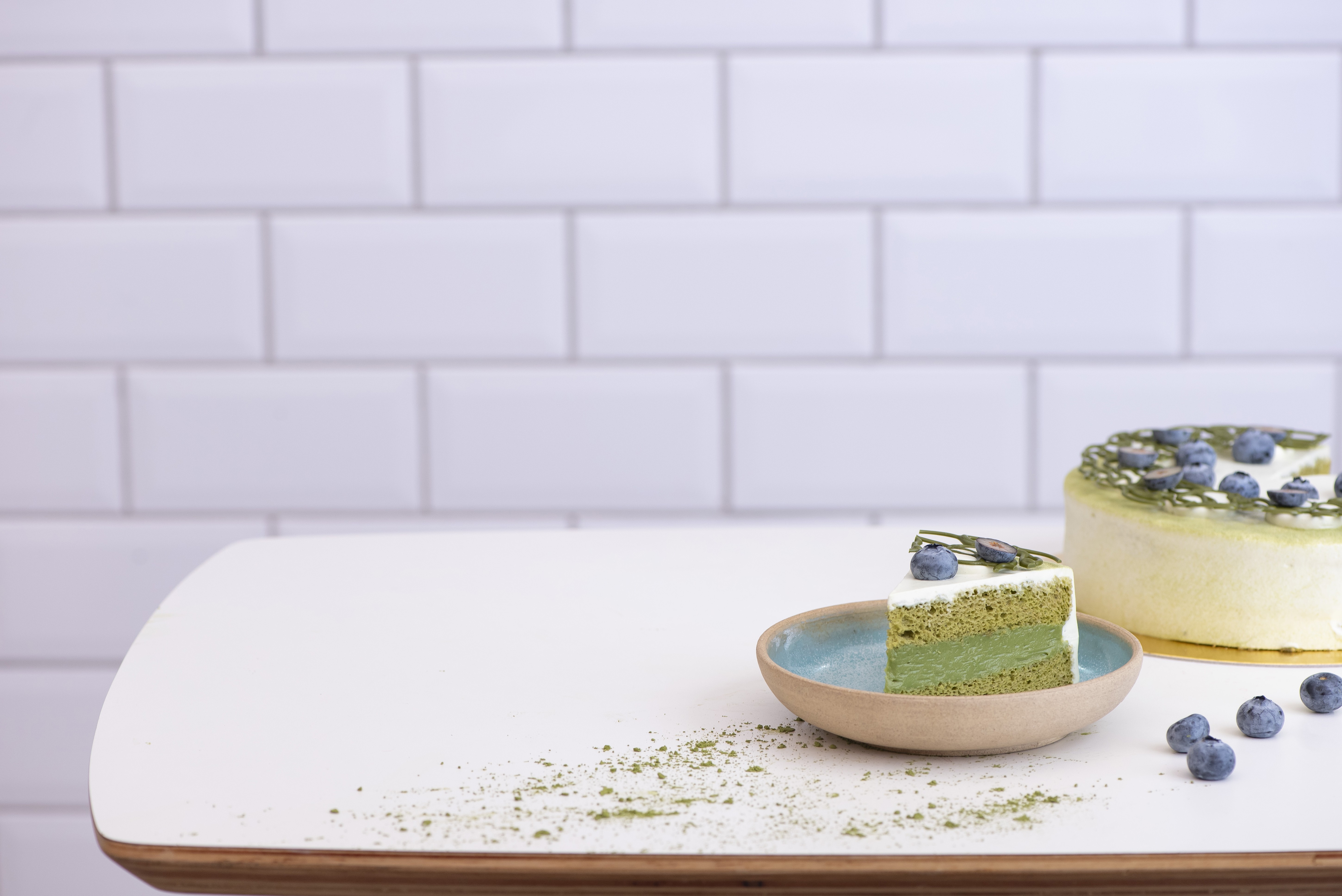 Fatia de bolo verde-claro com cobertura branca e mirtilos em um prato azul, com um bolo inteiro ao fundo, sobre uma mesa branca com fundo de azulejos brancos