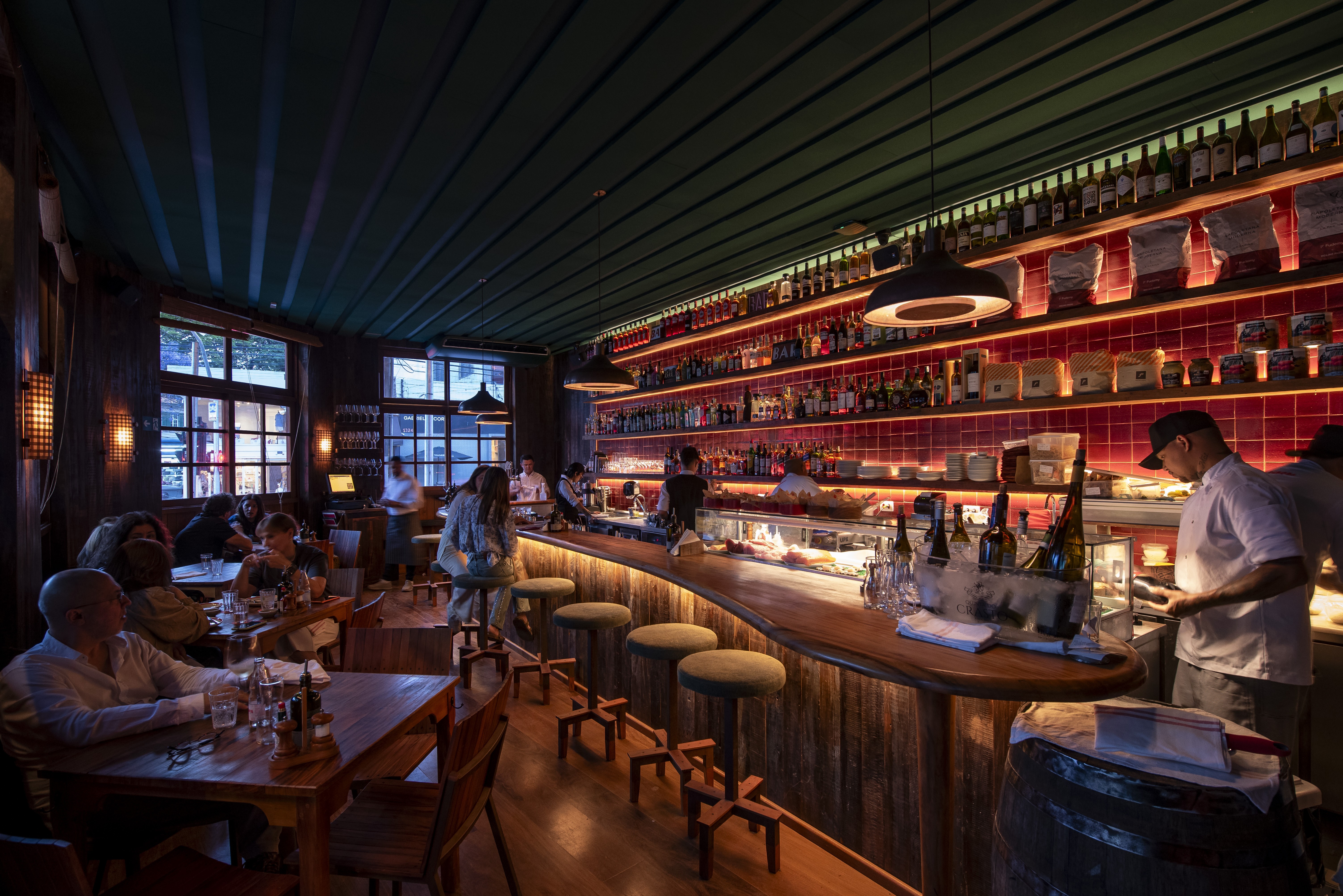 Interior de um bar ou restaurante com ilumina&ccedil;&atilde;o baixa e aconchegante. Clientes sentados em mesas de madeira e no balc&atilde;o, que tem banquetas de madeira e estofado verde. Atr&aacute;s do balc&atilde;o, prateleiras com garrafas e objetos decorativos, iluminadas por luzes vermelhas. Dois funcion&aacute;rios trabalham atr&aacute;s do balc&atilde;o, um deles de bon&eacute; e camisa branca. O teto &eacute; verde escuro com listras claras e h&aacute; janelas grandes ao fundo