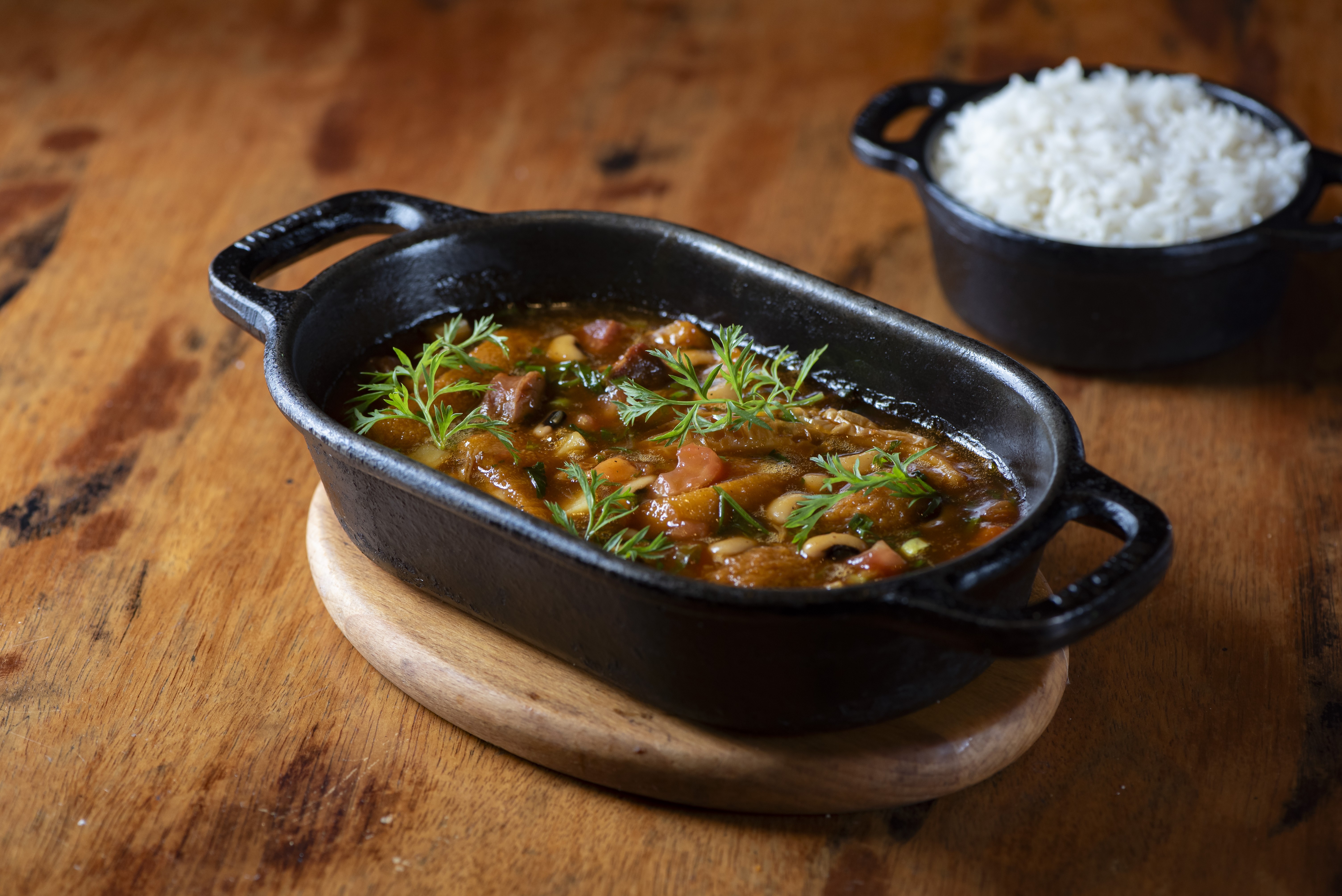 Ensopado de carne com legumes e feij&atilde;o-fradinho em panela de ferro preta, guarnecido com folhas verdes, sobre uma t&aacute;bua de madeira. Ao fundo, uma tigela de arroz branco