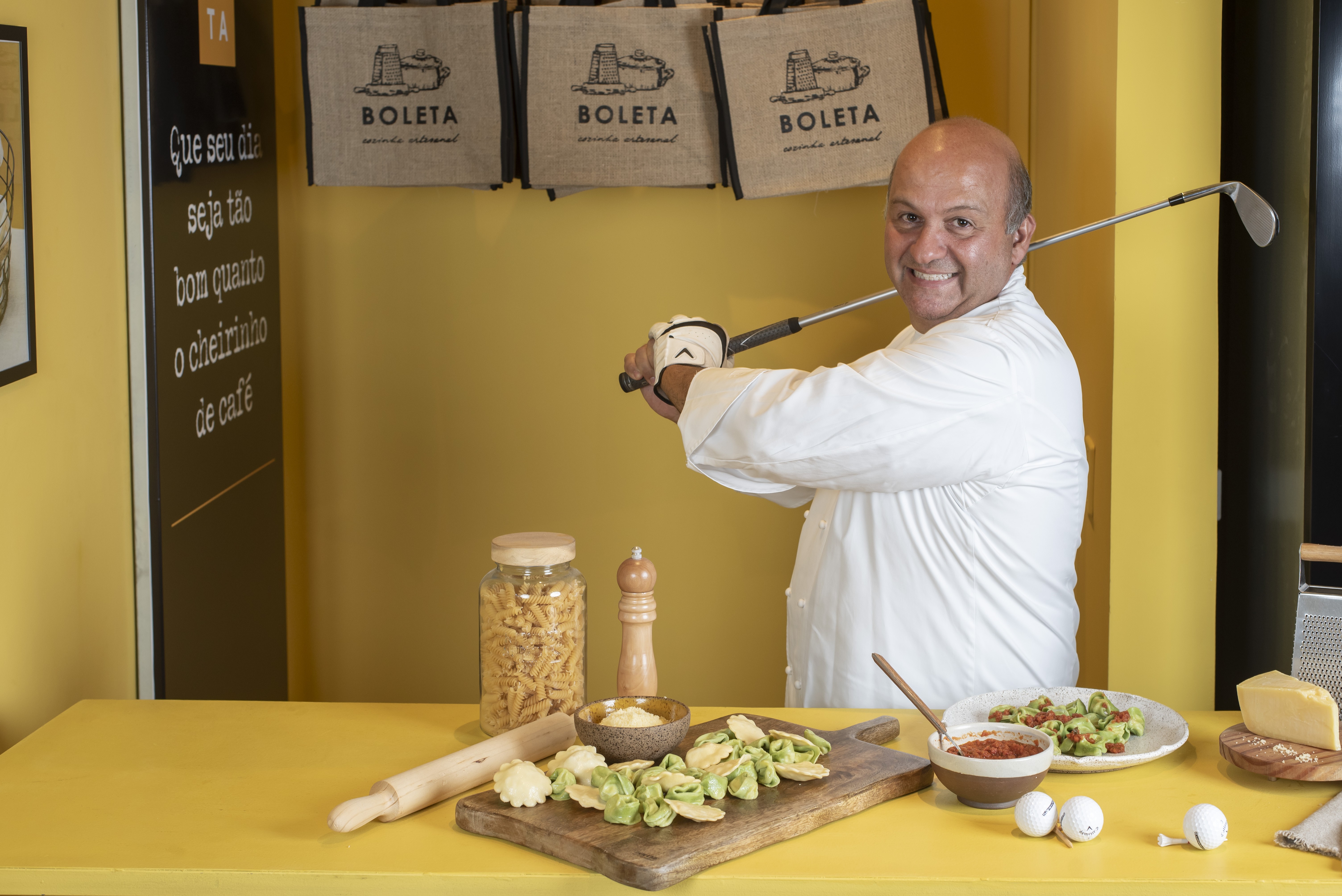 Homem careca, de camisa branca de chef, sorri segurando um taco de golfe sobre o ombro, em uma cozinha amarela com massas e queijo na bancada