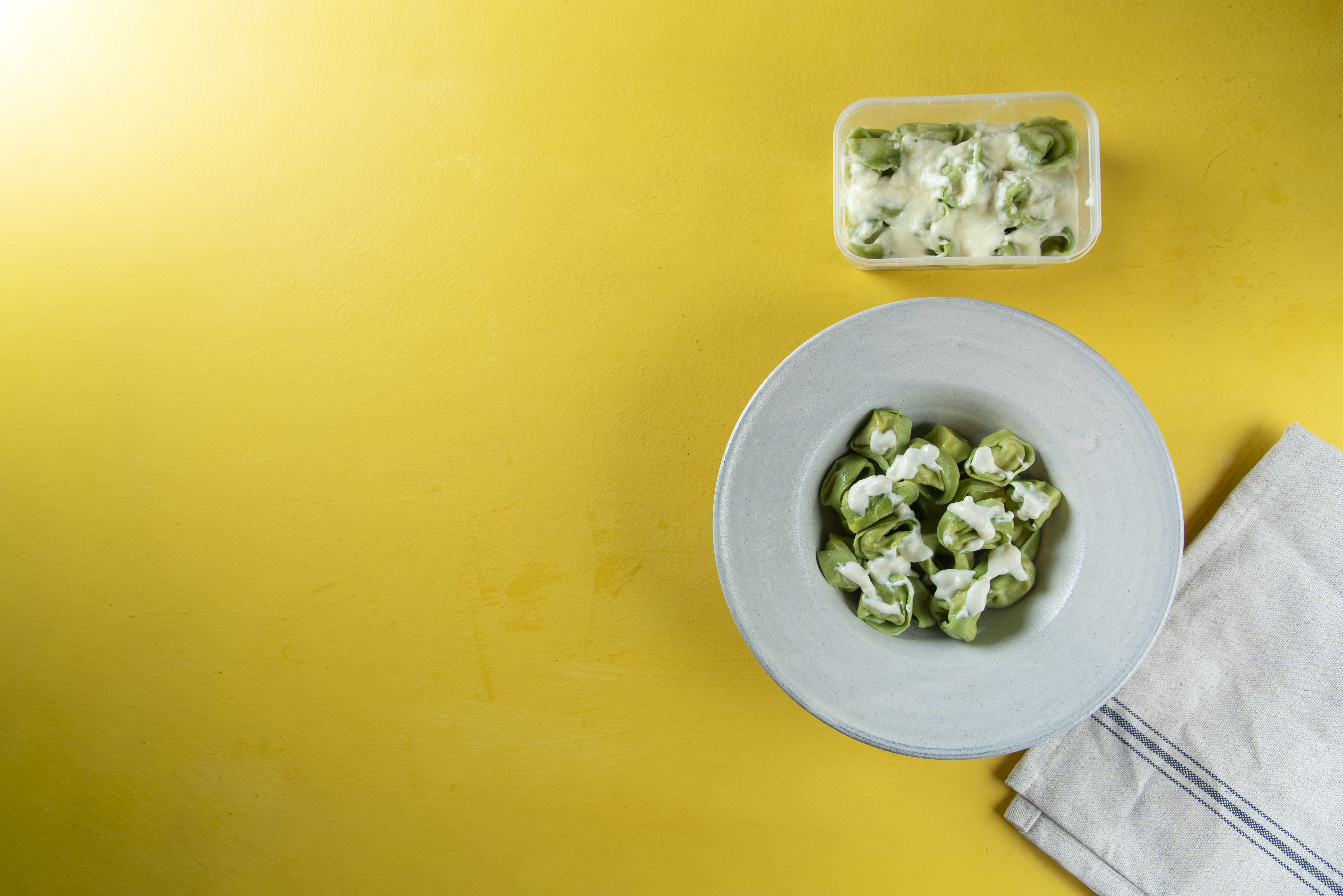 Prato de tortellini verde com molho branco cremoso, ao lado de um pote transparente com mais tortellini e um pano de prato dobrado, sobre uma superf&iacute;cie amarela