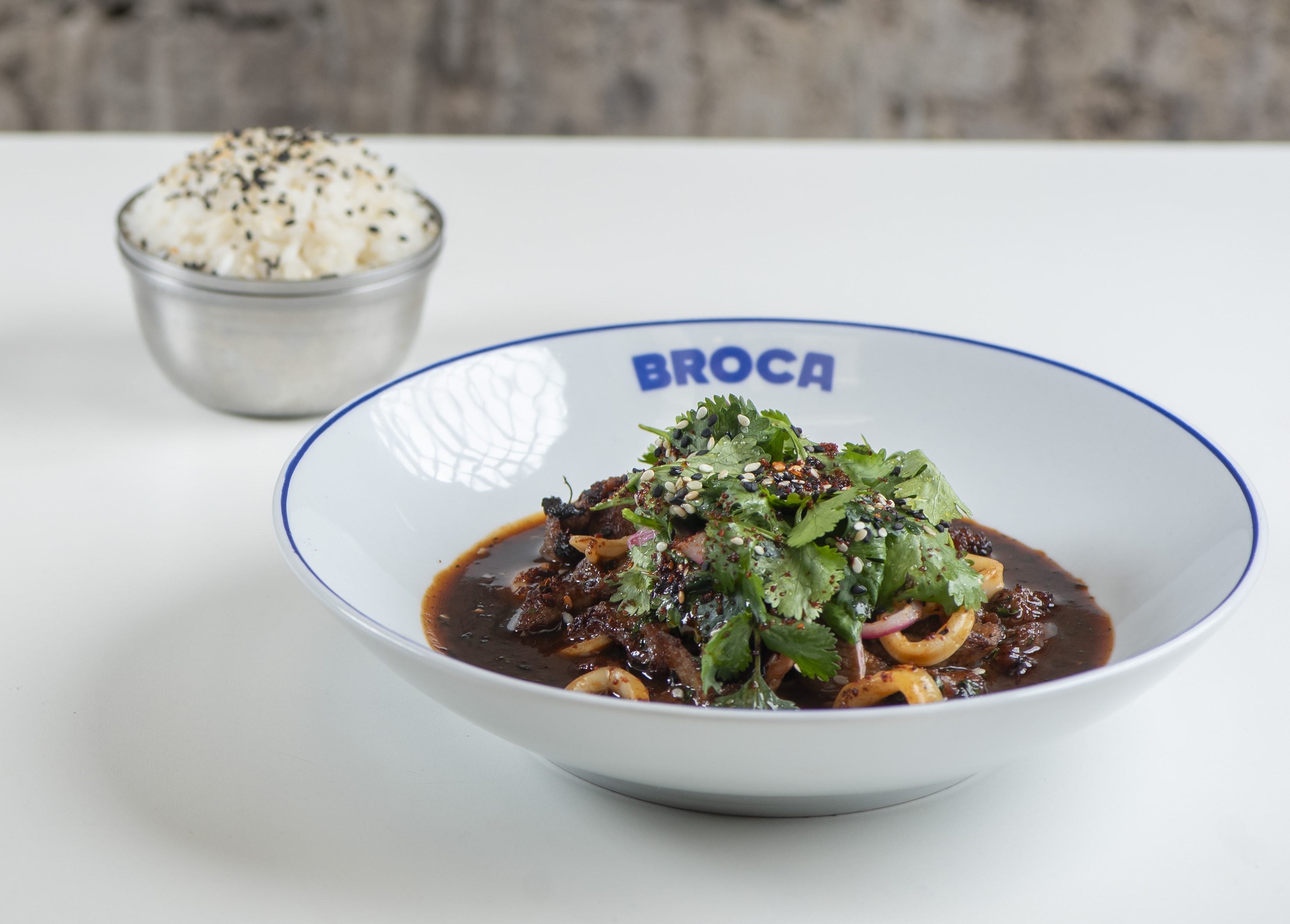 Prato branco com borda azul e nome BROCA em azul, contendo carne escura em molho, an&eacute;is de cebola roxa, coentro fresco e gergelim. Ao fundo, tigela de metal com arroz branco e gergelim preto