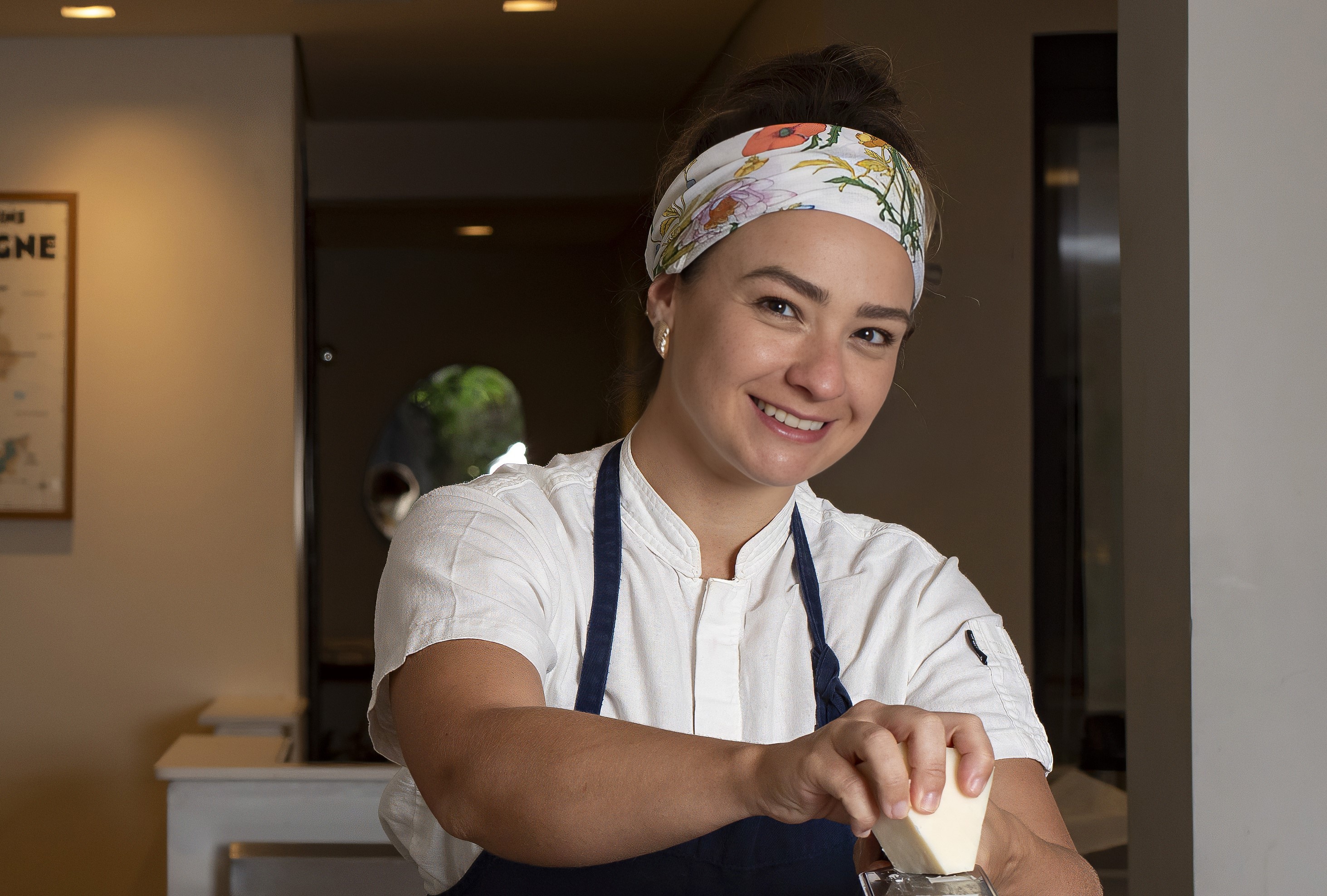 Cozinheira sorridente, de avental azul e bandana floral, ralando um peda&ccedil;o de queijo branco. Ela tem cabelos presos e brincos de p&eacute;rola, em um ambiente de cozinha com ilumina&ccedil;&atilde;o suave