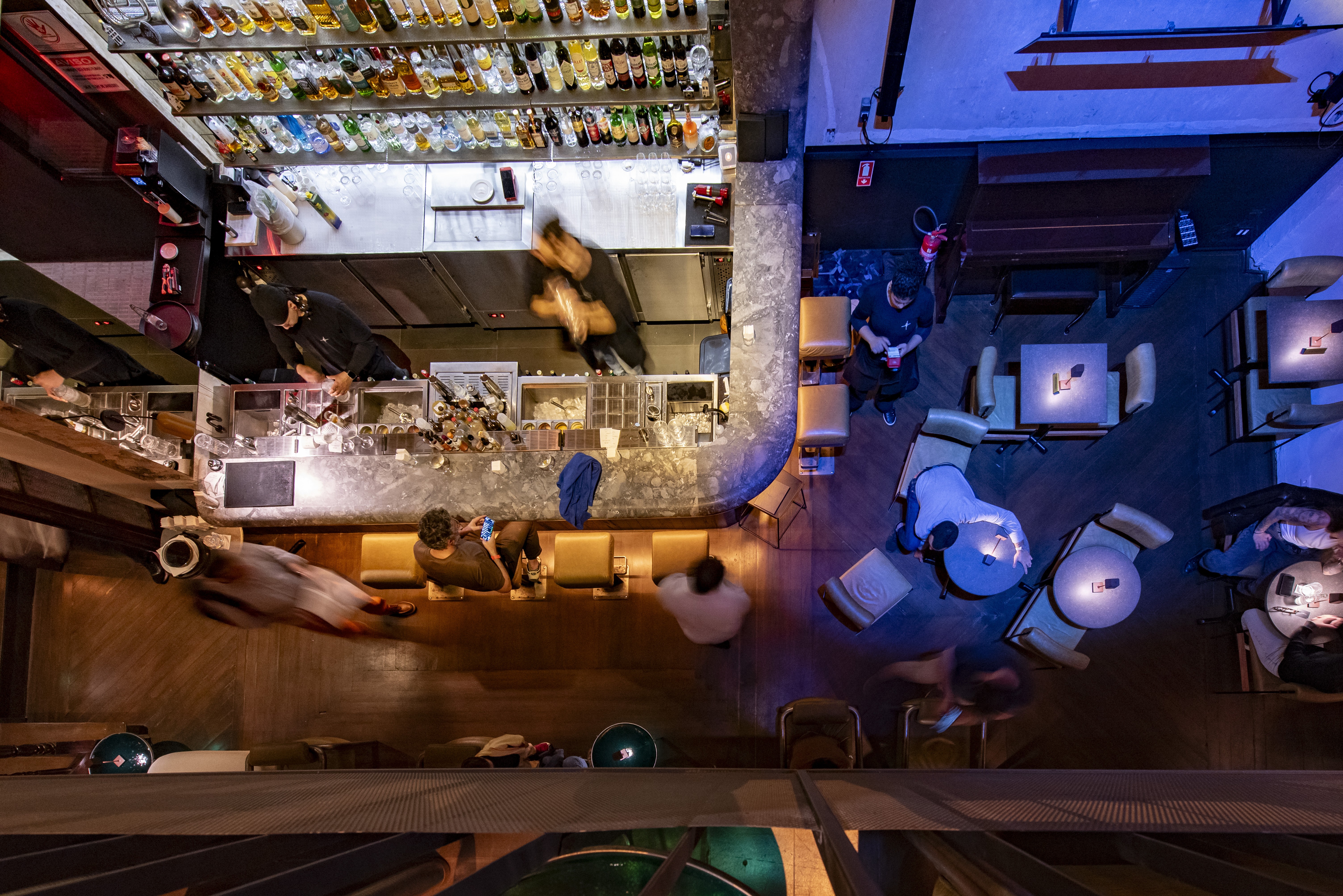 Vista a&eacute;rea de um bar movimentado com balc&atilde;o iluminado por cima, onde bartenders preparam bebidas. Clientes sentados em bancos no balc&atilde;o e em mesas redondas na &aacute;rea escura do sal&atilde;o, com luzes azuis e roxas. Algumas pessoas est&atilde;o borradas, indicando movimento