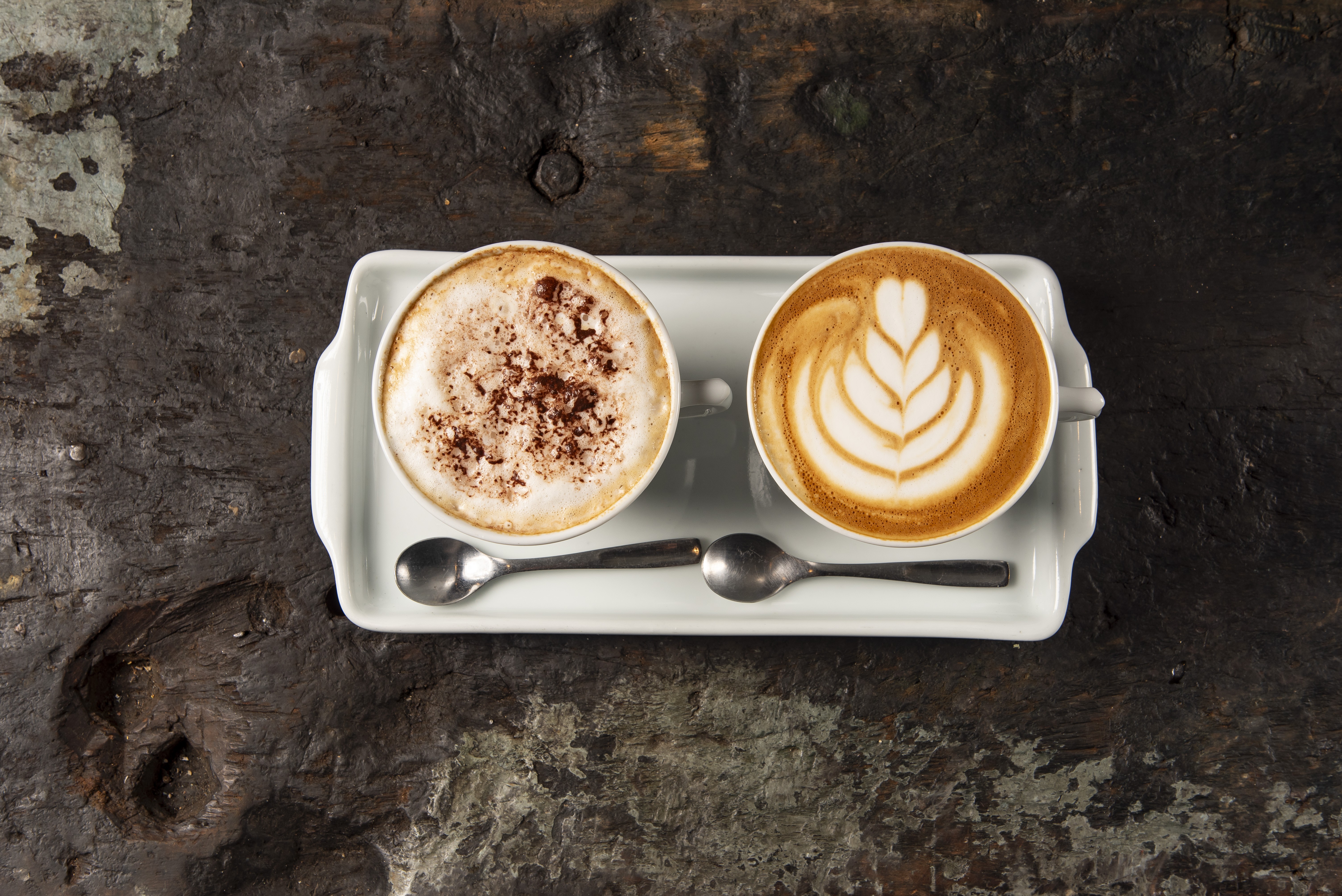 Duas x&iacute;caras de caf&eacute; em uma bandeja branca sobre uma mesa de madeira escura. A x&iacute;cara da esquerda tem espuma e chocolate em p&oacute;, a da direita tem um desenho de folha em latte art. Duas colheres de metal est&atilde;o entre as x&iacute;caras