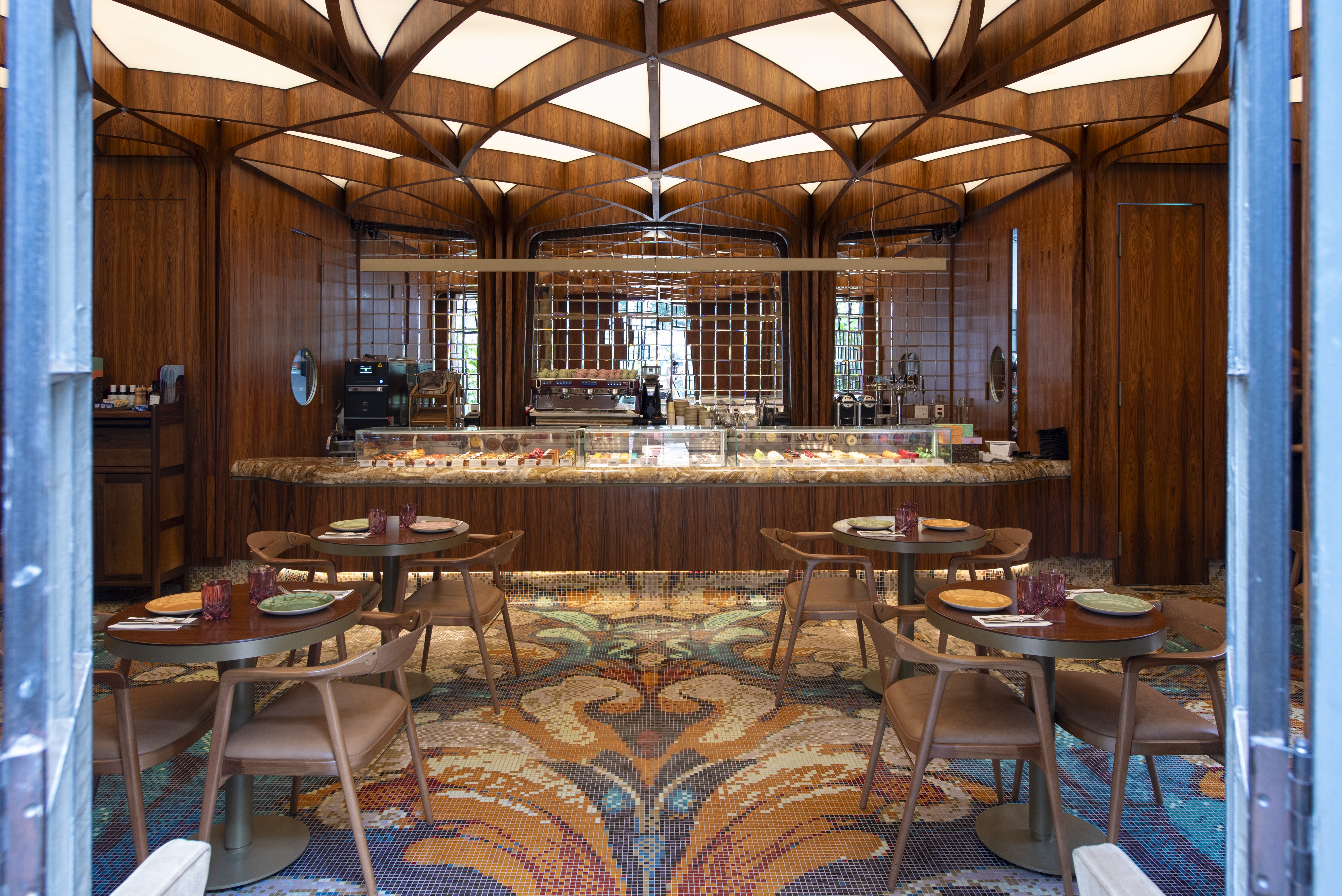 Interior de um caf&eacute; ou restaurante luxuoso com teto abobadado de madeira e ilumina&ccedil;&atilde;o embutida. O balc&atilde;o principal exibe doces e salgados. Mesas redondas de madeira com cadeiras estofadas em couro marrom est&atilde;o dispostas sobre um piso de mosaico colorido com padr&otilde;es florais e geom&eacute;tricos. As paredes s&atilde;o de madeira escura, e janelas gradeadas refletem a luz externa