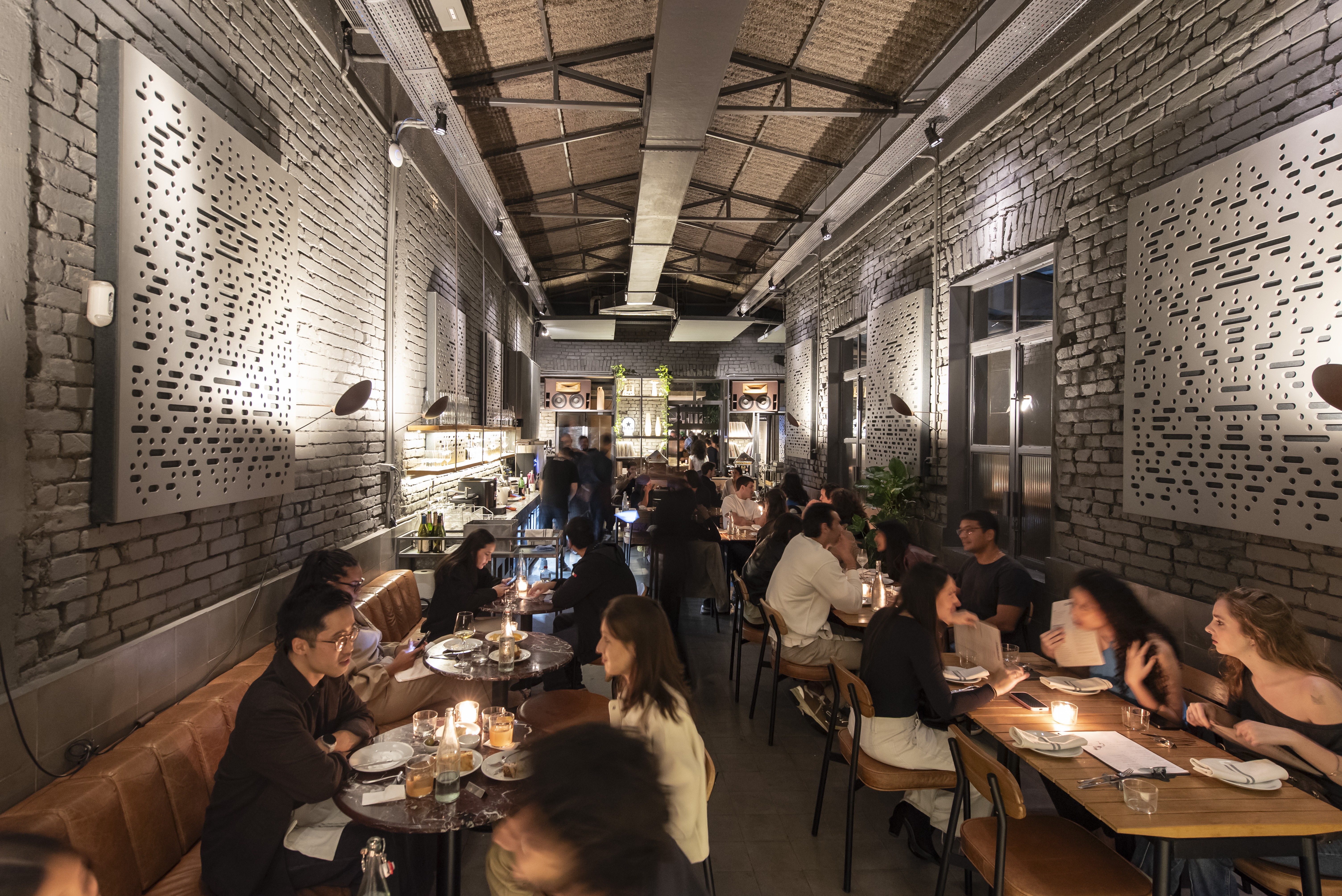 Interior de um restaurante moderno com paredes de tijolos aparentes e teto alto. V&aacute;rias pessoas sentadas em mesas e sof&aacute;s de couro marrom, conversando e jantando &agrave; luz de velas. Pain&eacute;is decorativos brancos com recortes geom&eacute;tricos adornam as paredes, iluminados por spots. Ao fundo, uma &aacute;rea de bar com garrafas e utens&iacute;lios. O ambiente &eacute; aconchegante e movimentado