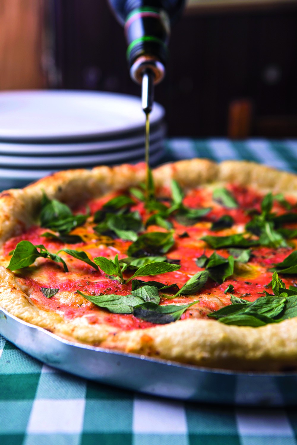 Pizza margherita rec&eacute;m-assada em forma de metal, com molho de tomate e folhas de manjeric&atilde;o fresco, recebendo um fio de azeite de uma garrafa escura. Ao fundo, pratos brancos empilhados sobre uma toalha de mesa xadrez verde e branca