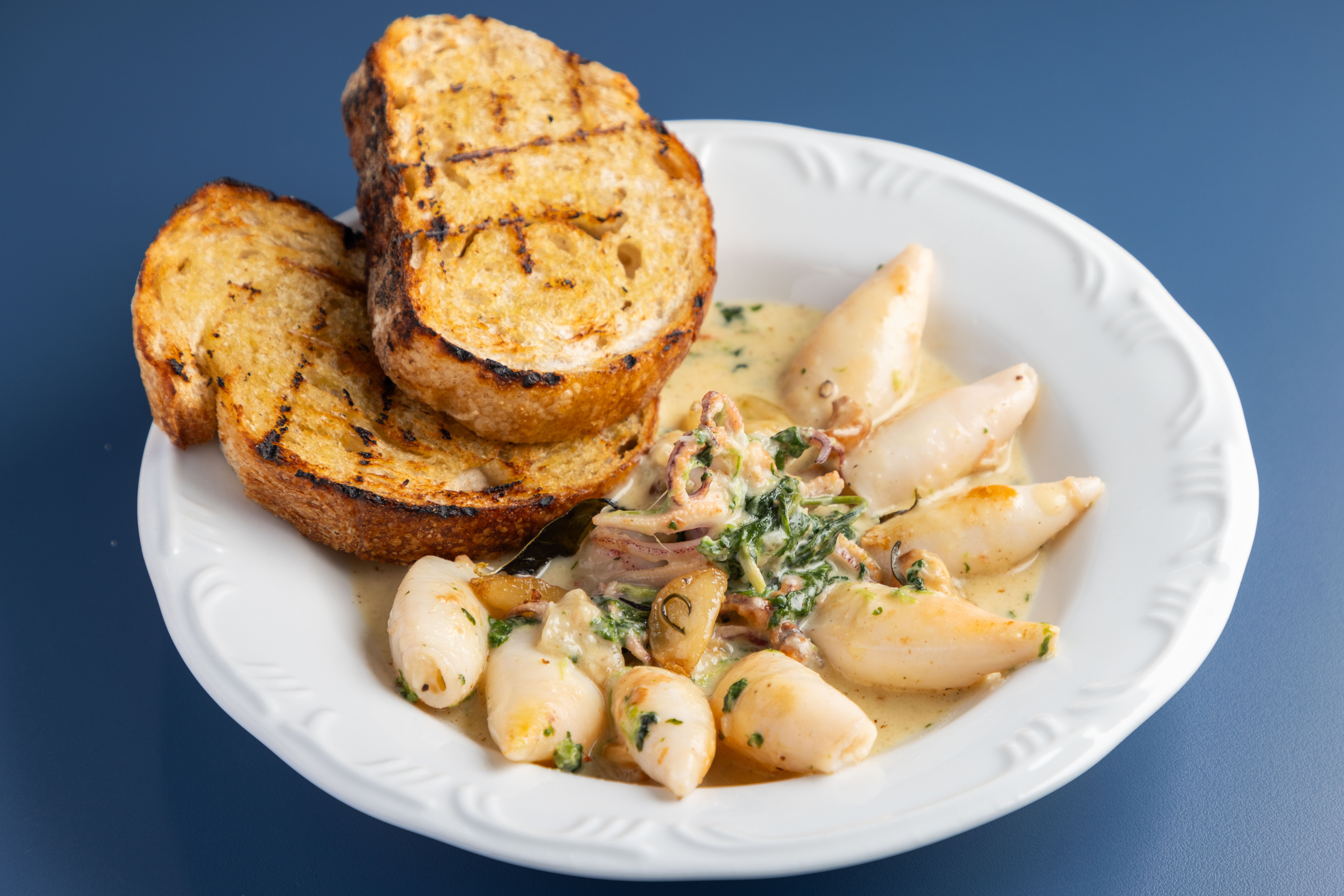 Lulas recheadas com molho cremoso de espinafre e bacon, servidas com duas fatias de p&atilde;o italiano grelhado, em um prato branco sobre fundo azul