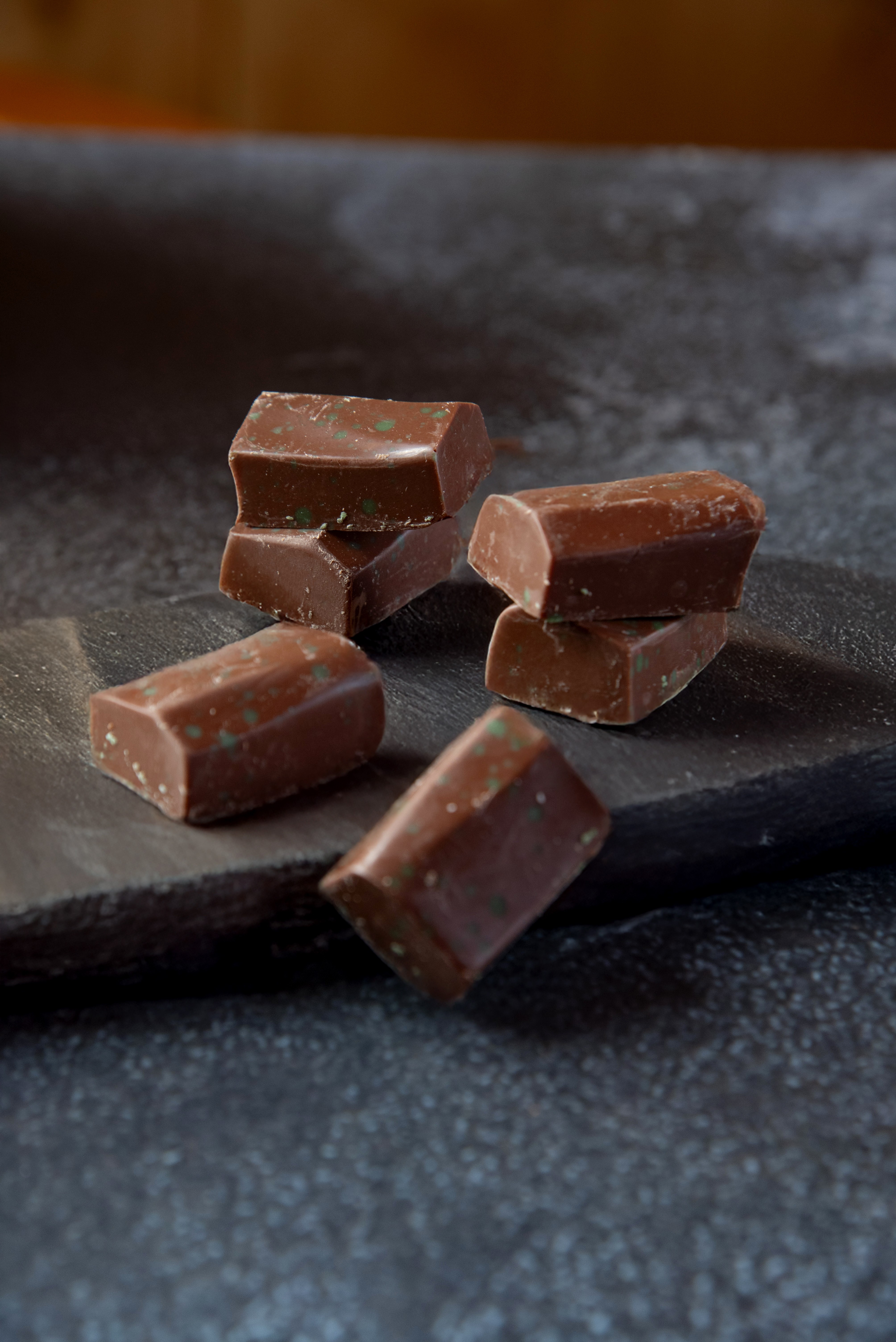 Seis bombons de chocolate ao leite, com pontos verdes e brancos, dispostos sobre uma t&aacute;bua de madeira escura. Dois est&atilde;o empilhados &agrave; esquerda, dois &agrave; direita e os outros dois espalhados na frente. O fundo &eacute; escuro e desfocado