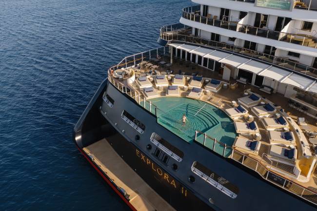 Vista a&eacute;rea de um navio de cruzeiro de luxo, o Explora II, com uma pessoa nadando na piscina do conv&eacute;s principal. O conv&eacute;s possui espregui&ccedil;adeiras e &aacute;reas de lounge, e o navio est&aacute; em &aacute;guas azuis claras sob um c&eacute;u ensolarado
