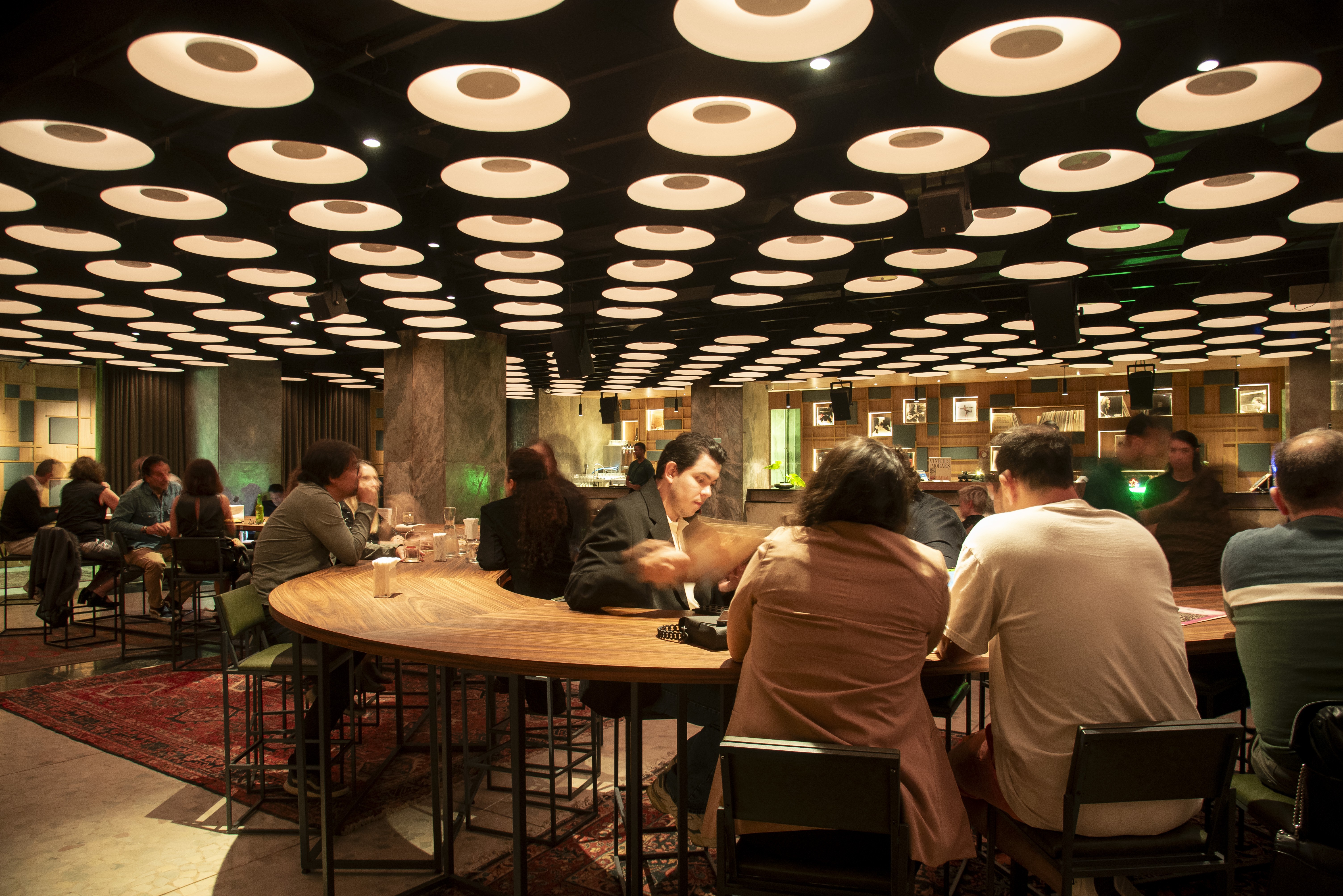 Pessoas sentadas em mesas de madeira em um restaurante ou bar com ilumina&ccedil;&atilde;o circular no teto, conversando e interagindo
