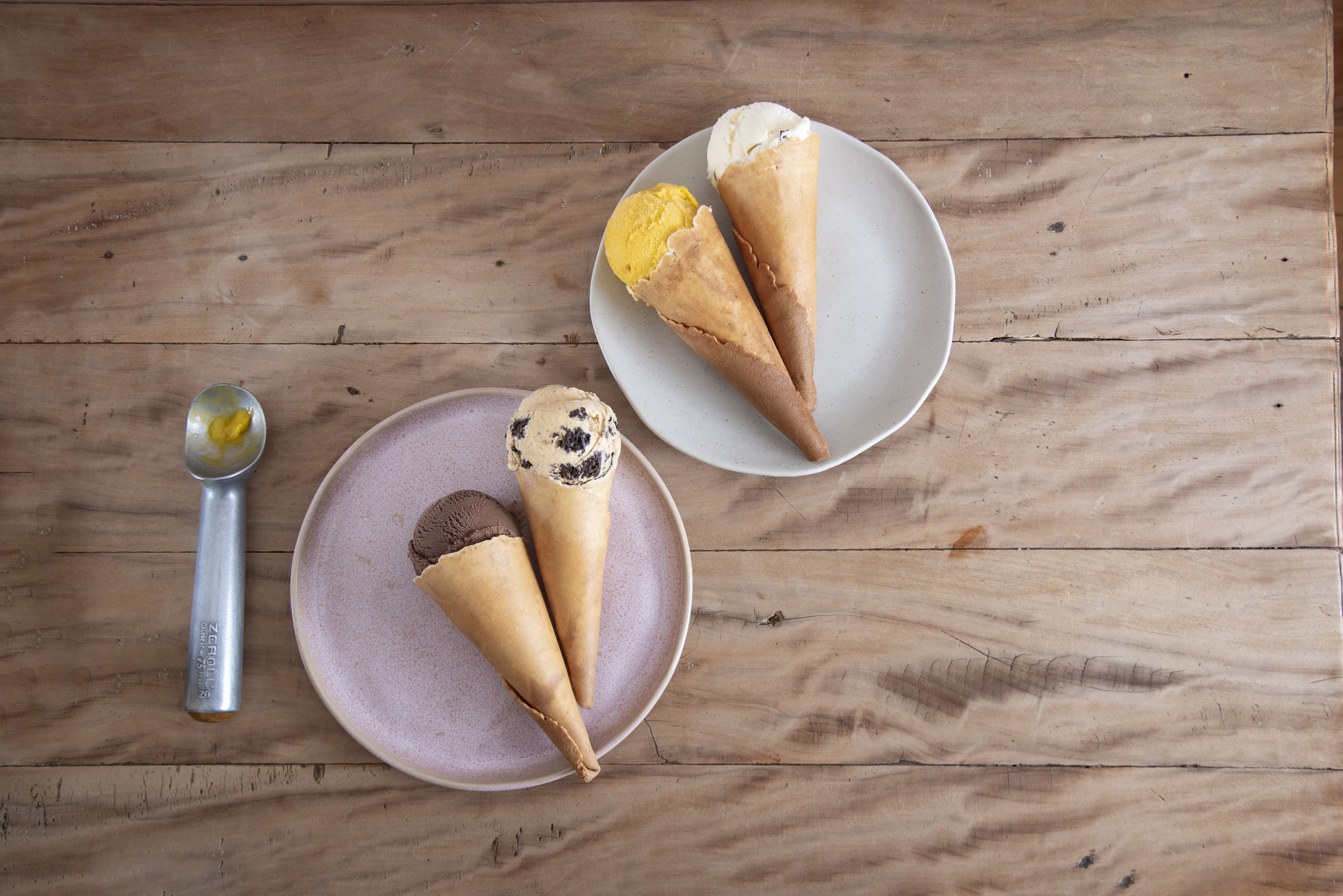 Quatro casquinhas de sorvete em pratos redondos sobre uma mesa de madeira clara. No prato superior, sorvetes de baunilha e manga. No prato inferior, sorvetes de chocolate e creme com peda&ccedil;os de biscoito. Uma colher de sorvete met&aacute;lica est&aacute; &agrave; esquerda