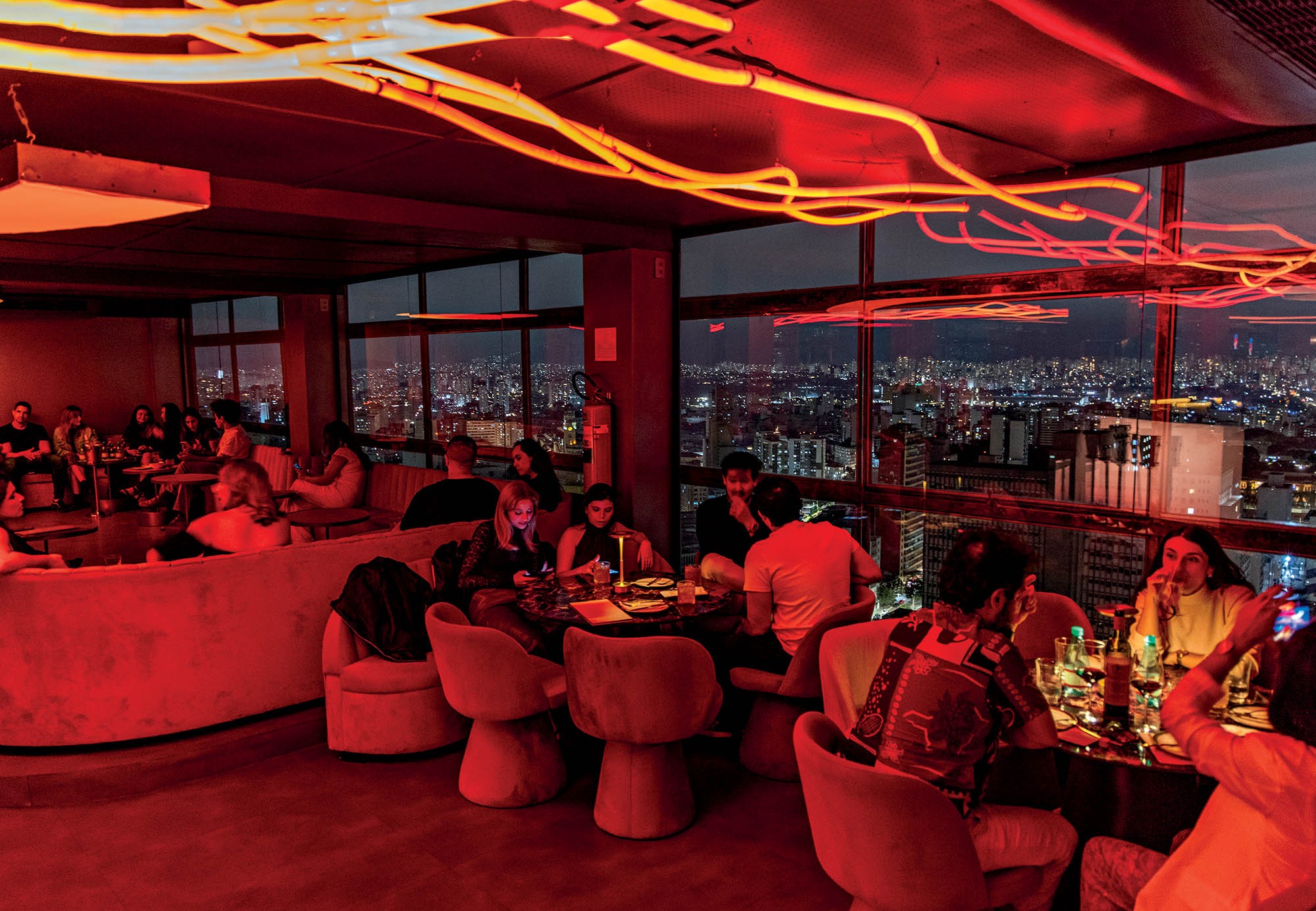 Pessoas em um bar com iluminação vermelha e neon laranja, com vista panorâmica de uma cidade à noite. Grupos conversam em mesas e sofás, alguns com bebidas, em um ambiente animado e moderno