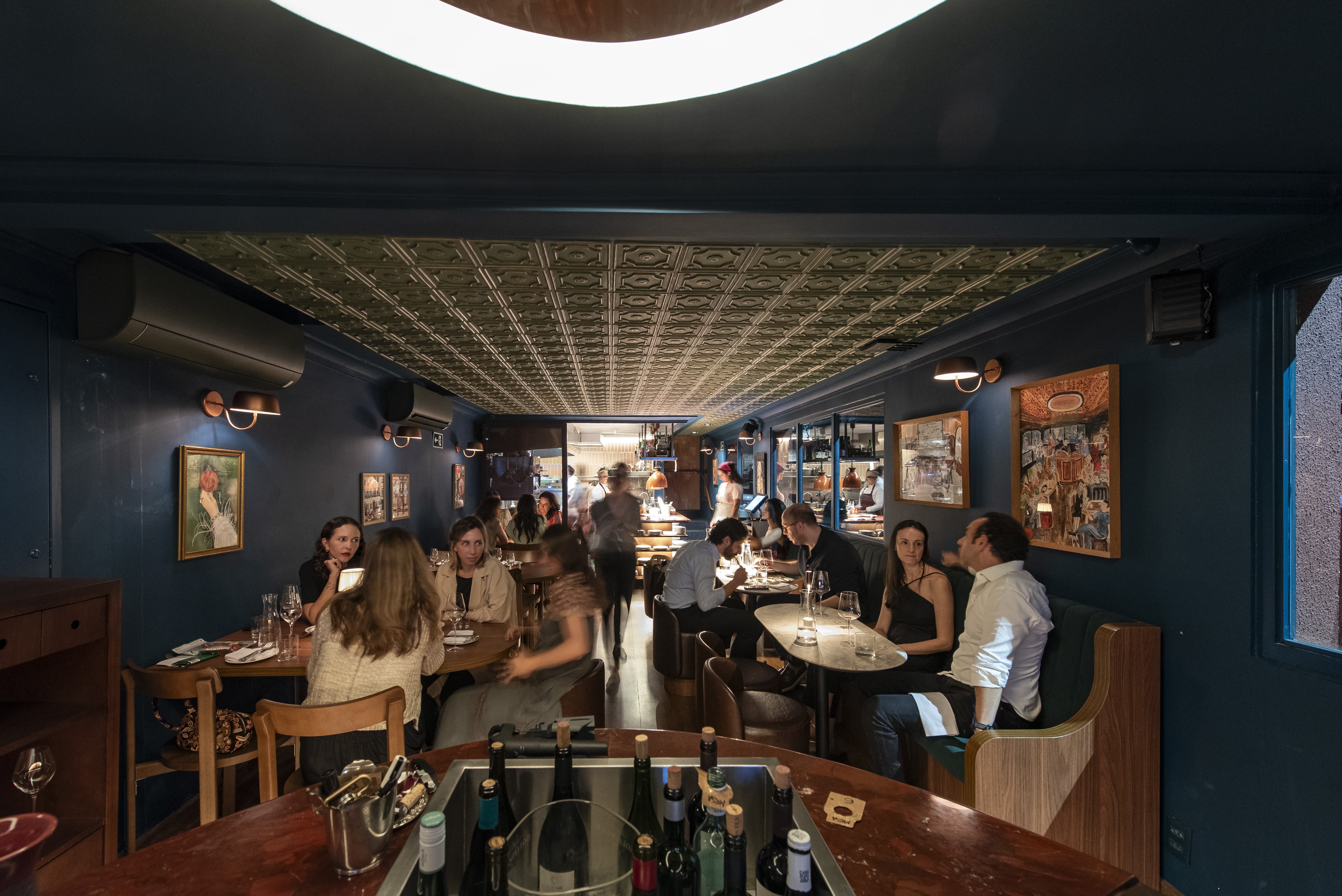 Pessoas conversam em um restaurante com paredes azul-marinho e teto de metal estampado. Mesas redondas de madeira e cadeiras estofadas marrons ocupam o espa&ccedil;o, com garrafas de vinho em um balc&atilde;o em primeiro plano. Quadros decoram as paredes, e a ilumina&ccedil;&atilde;o &eacute; suave, criando um ambiente acolhedor