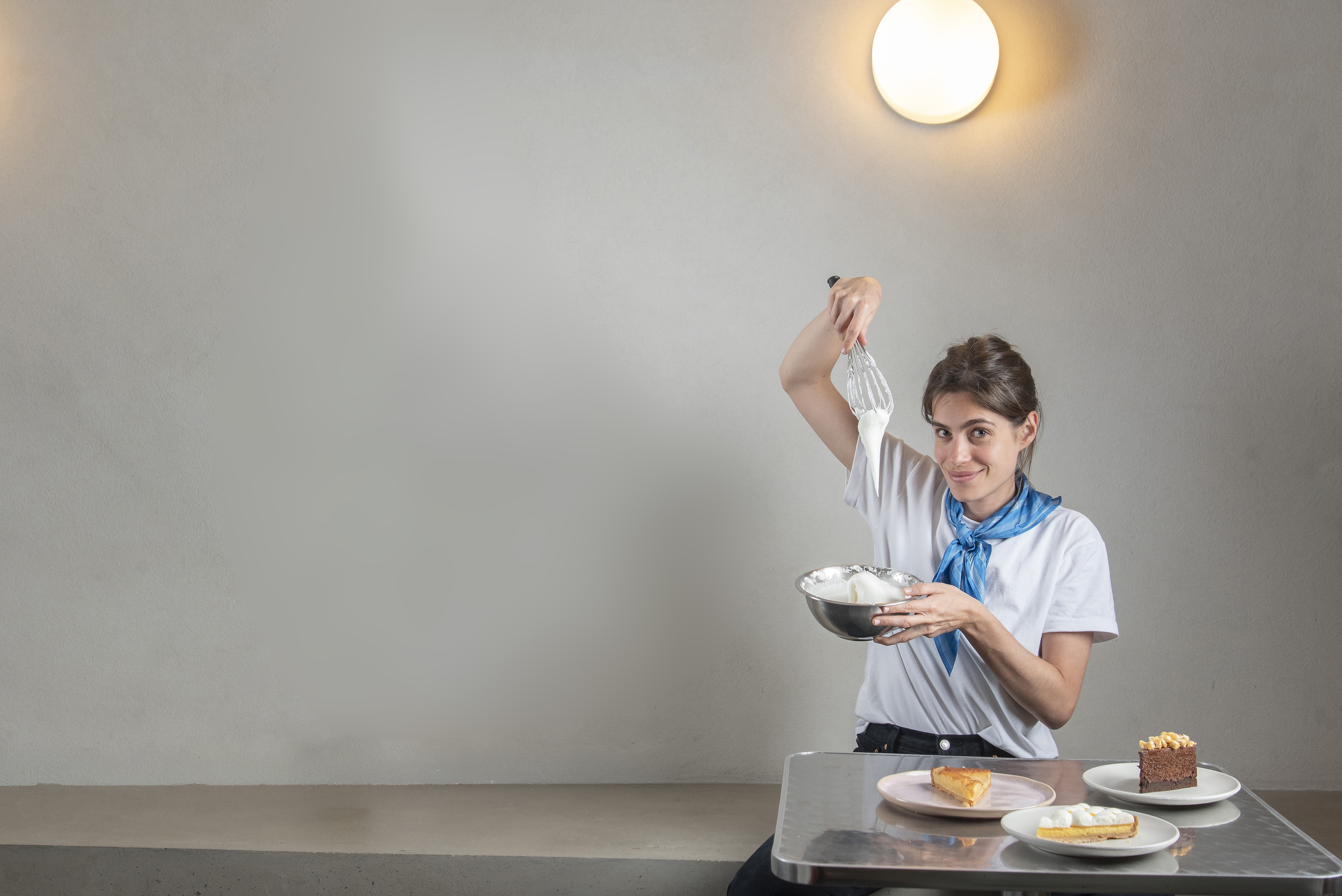 Mulher jovem sorridente, vestindo camiseta branca e len&ccedil;o azul, segura um batedor com creme e uma tigela com chantilly, sentada &agrave; mesa com fatias de torta e bolo