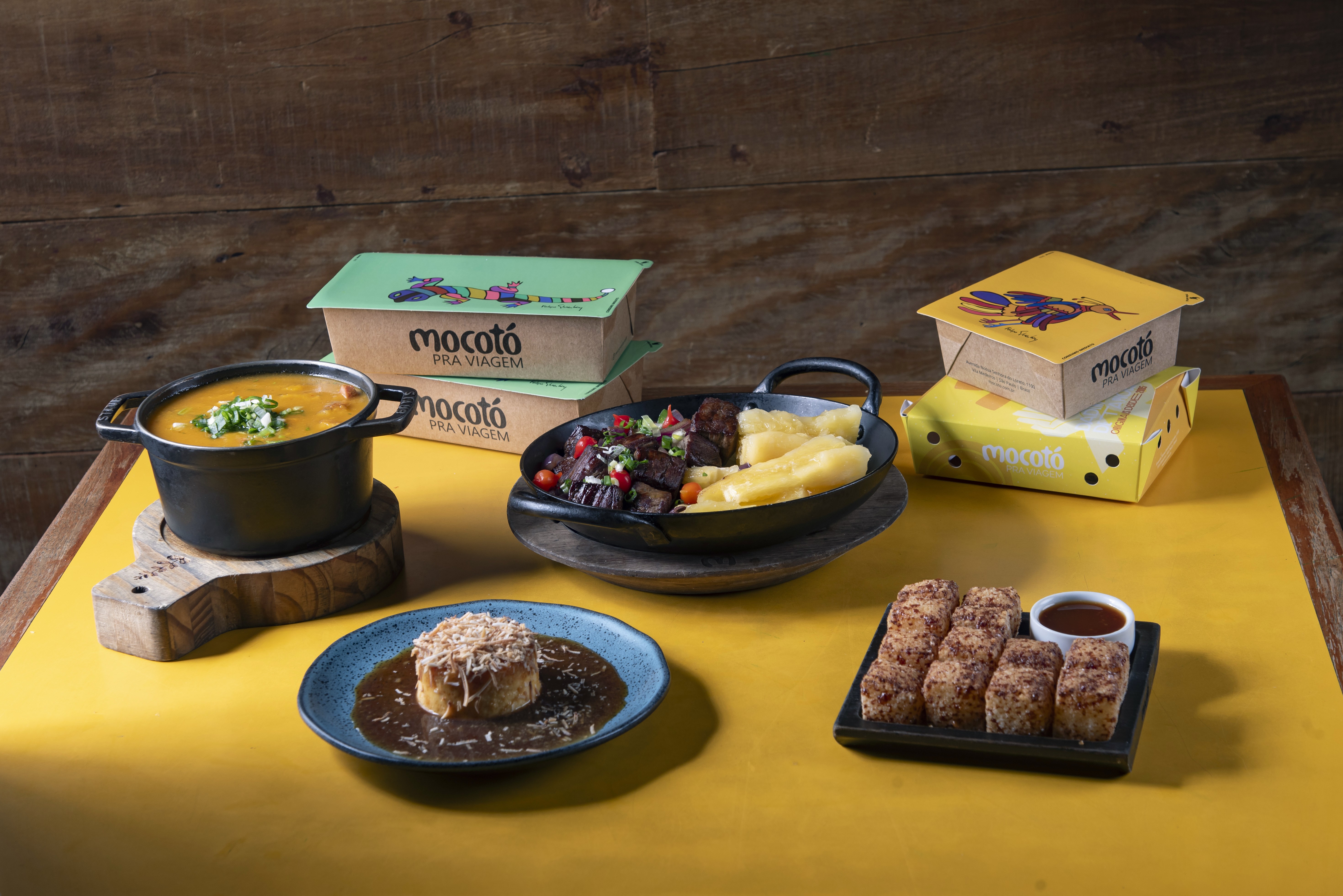 Mesa com pratos do Mocot&oacute;: uma panela de feij&atilde;o, uma frigideira com carne e mandioca, um bolinho com molho, por&ccedil;&otilde;es de dadinhos de tapioca e embalagens para viagem, sobre um fundo amarelo e madeira