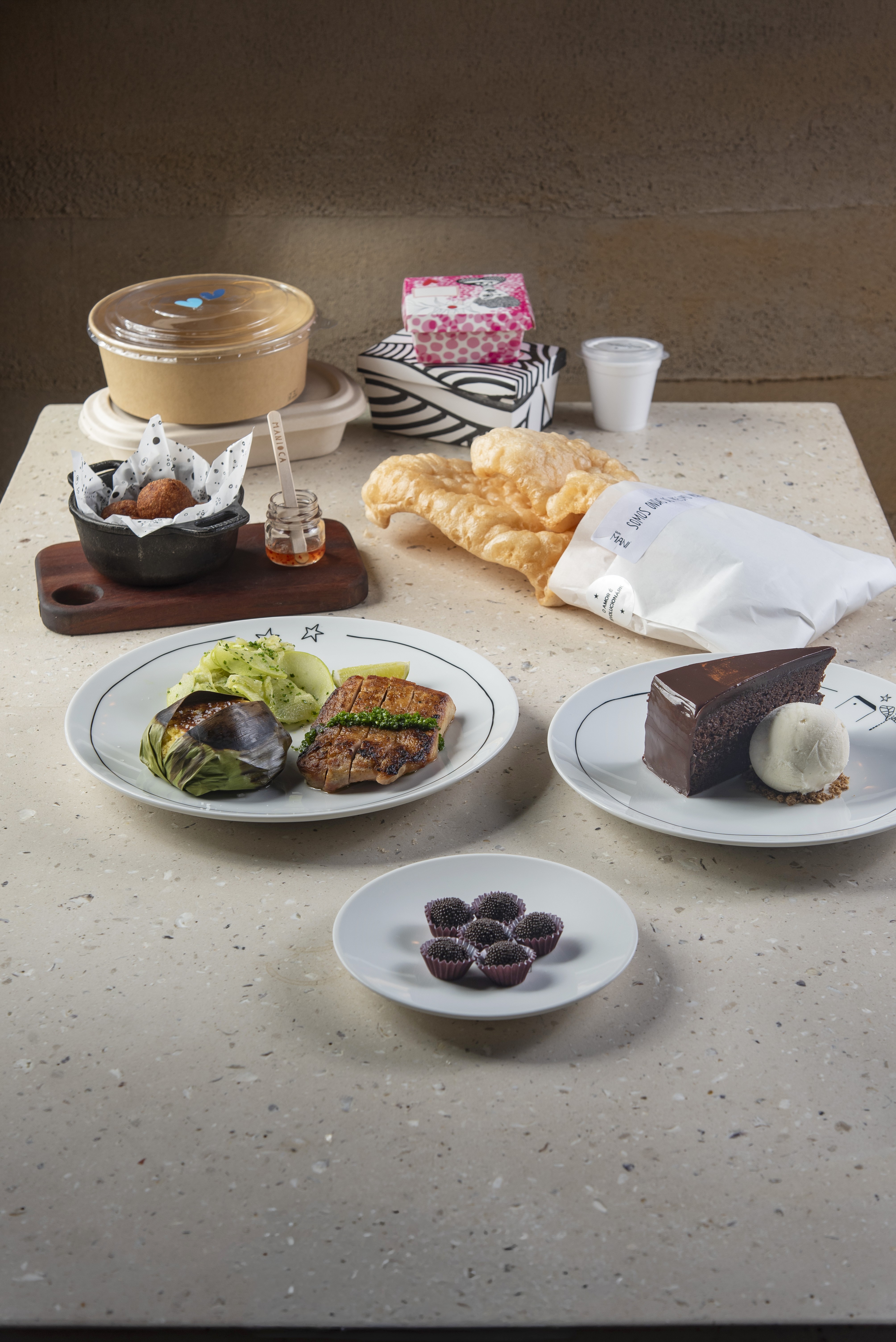 Mesa com refei&ccedil;&atilde;o completa: prato principal com peixe grelhado e pur&ecirc;, bolo de chocolate com sorvete, brigadeiros, por&ccedil;&otilde;es de torresmo e bolinhos, e embalagens de delivery
