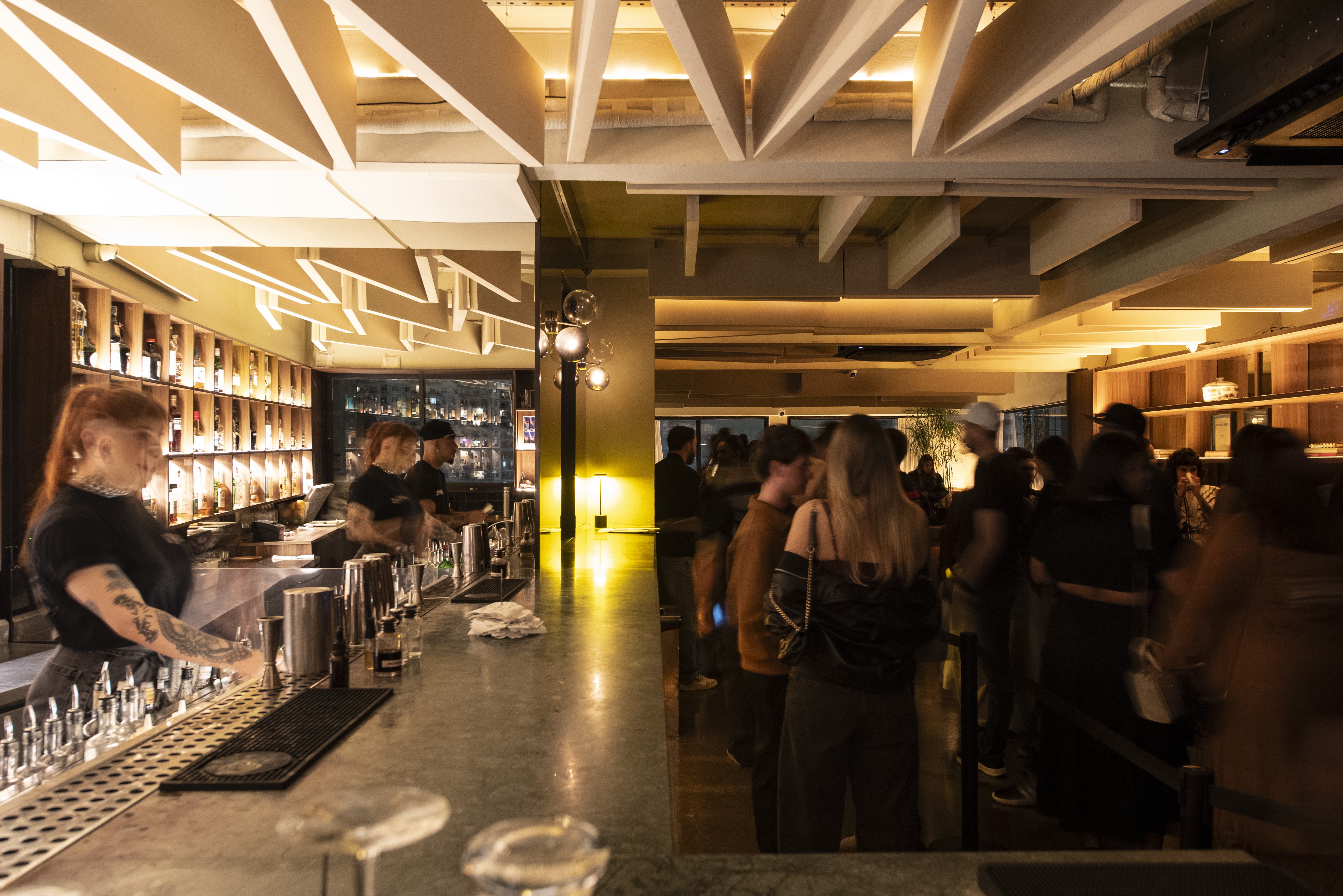 Mulher ruiva tatuada com blusa preta serve bebidas em um bar movimentado com teto de madeira e luzes quentes, enquanto clientes conversam e esperam