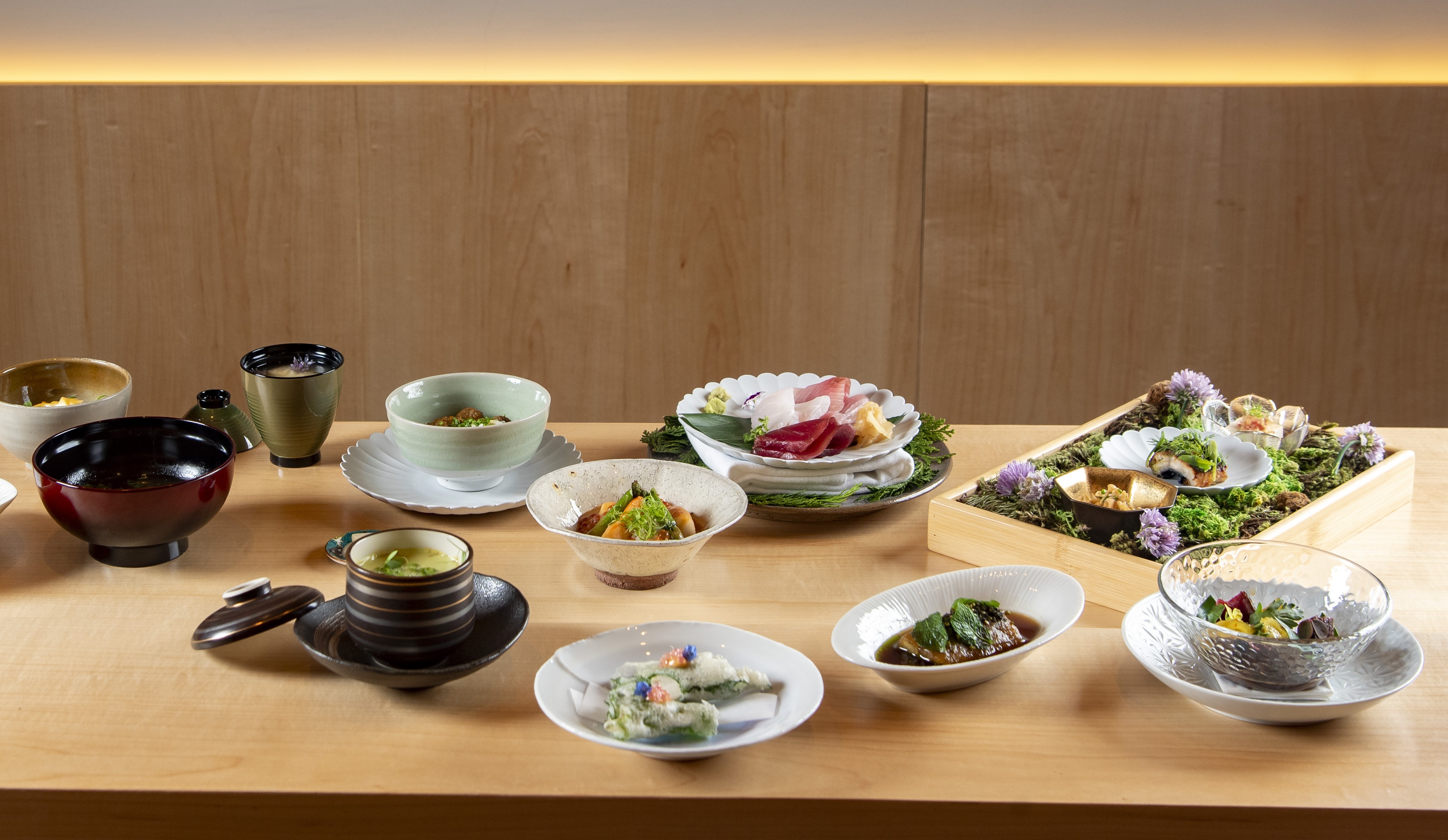 Mesa de madeira clara com uma variedade de pratos japoneses em lou&ccedil;as e tigelas de diferentes cores e tamanhos, incluindo sashimi, sopas, saladas e um arranjo de musgo com pequenas por&ccedil;&otilde;es de comida e flores roxas