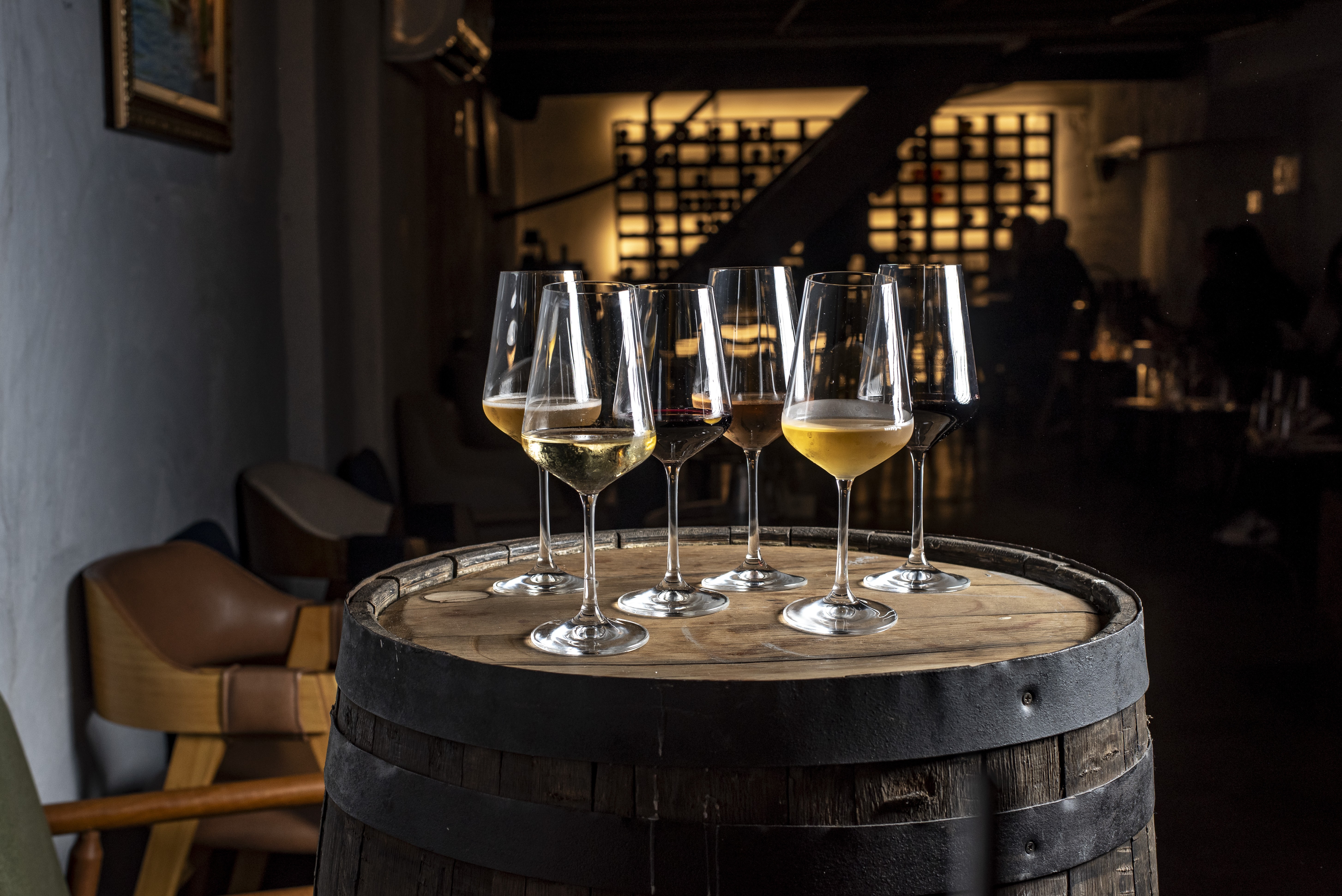 Seis ta&ccedil;as de vinho com diferentes tonalidades de branco, ros&eacute; e tinto sobre um barril de madeira r&uacute;stico, em um ambiente escuro de adega com prateleiras de garrafas ao fundo