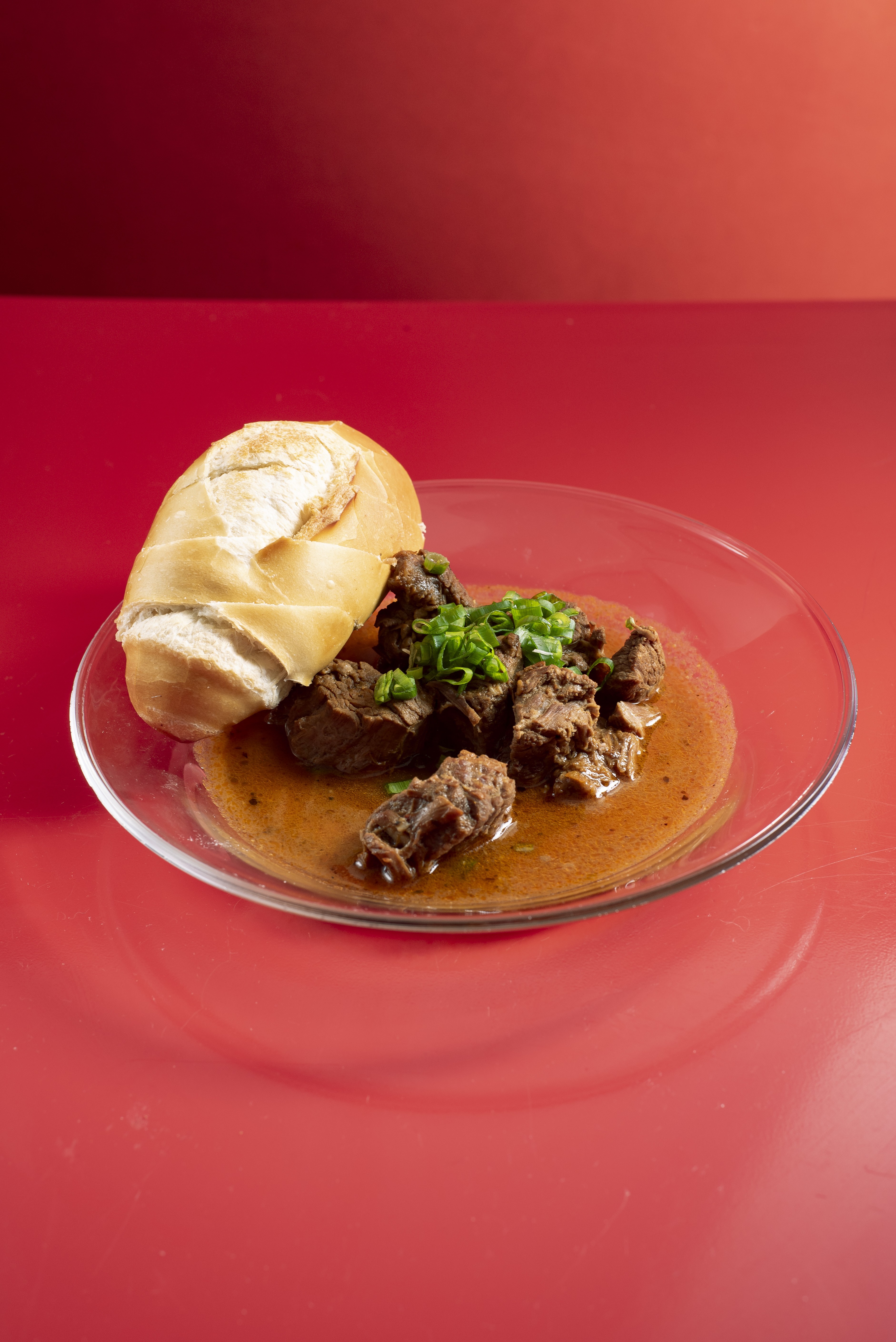 Um prato de vidro transparente com carne de panela em molho marrom-avermelhado, guarnecida com cebolinha picada, e um p&atilde;o franc&ecirc;s cortado ao meio, sobre uma superf&iacute;cie vermelha brilhante