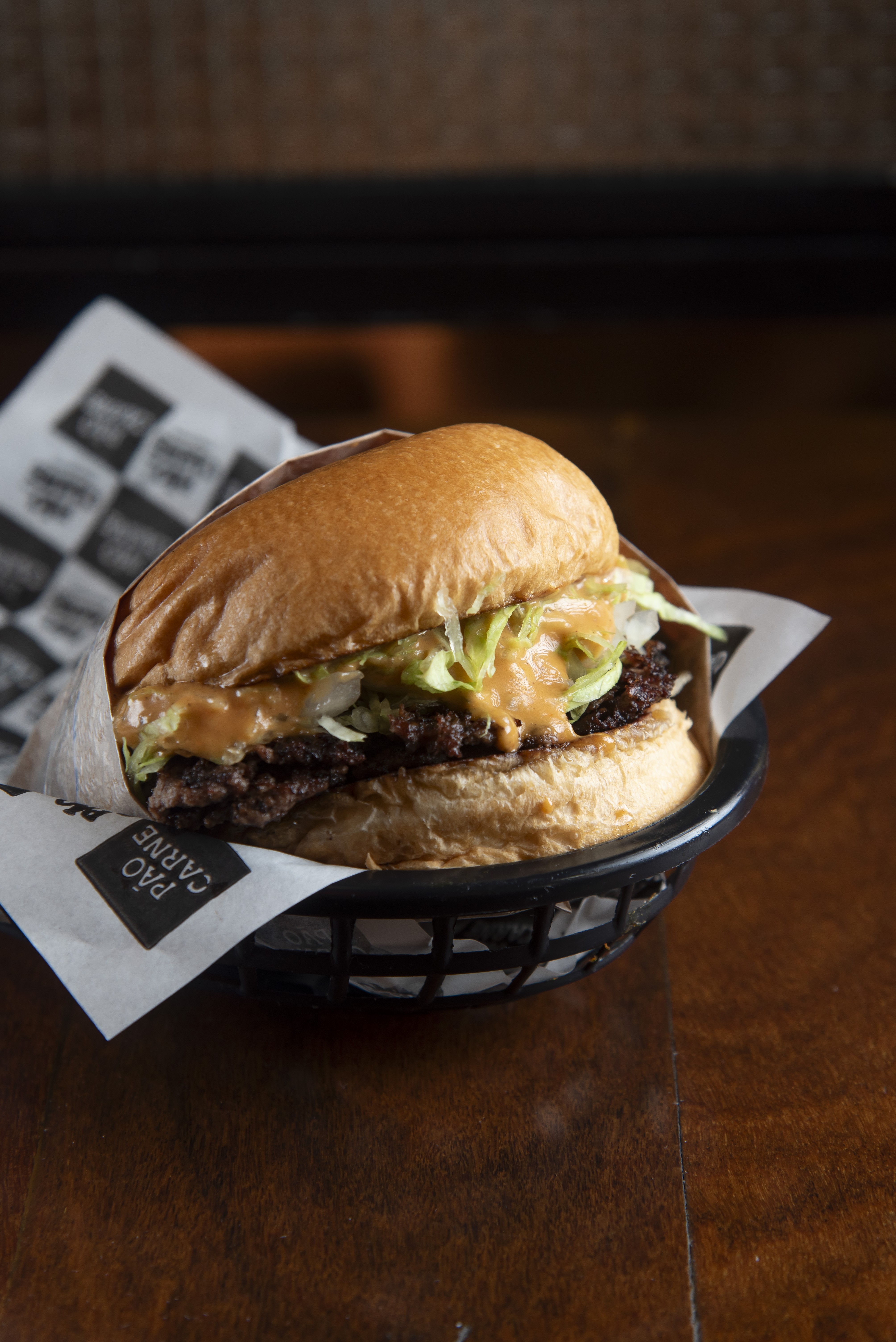 Hamb&uacute;rguer com dois bifes de carne, alface, cebola e molho cremoso, servido em cesto preto com papel estampado P&Atilde;O CARNE sobre mesa de madeira escura