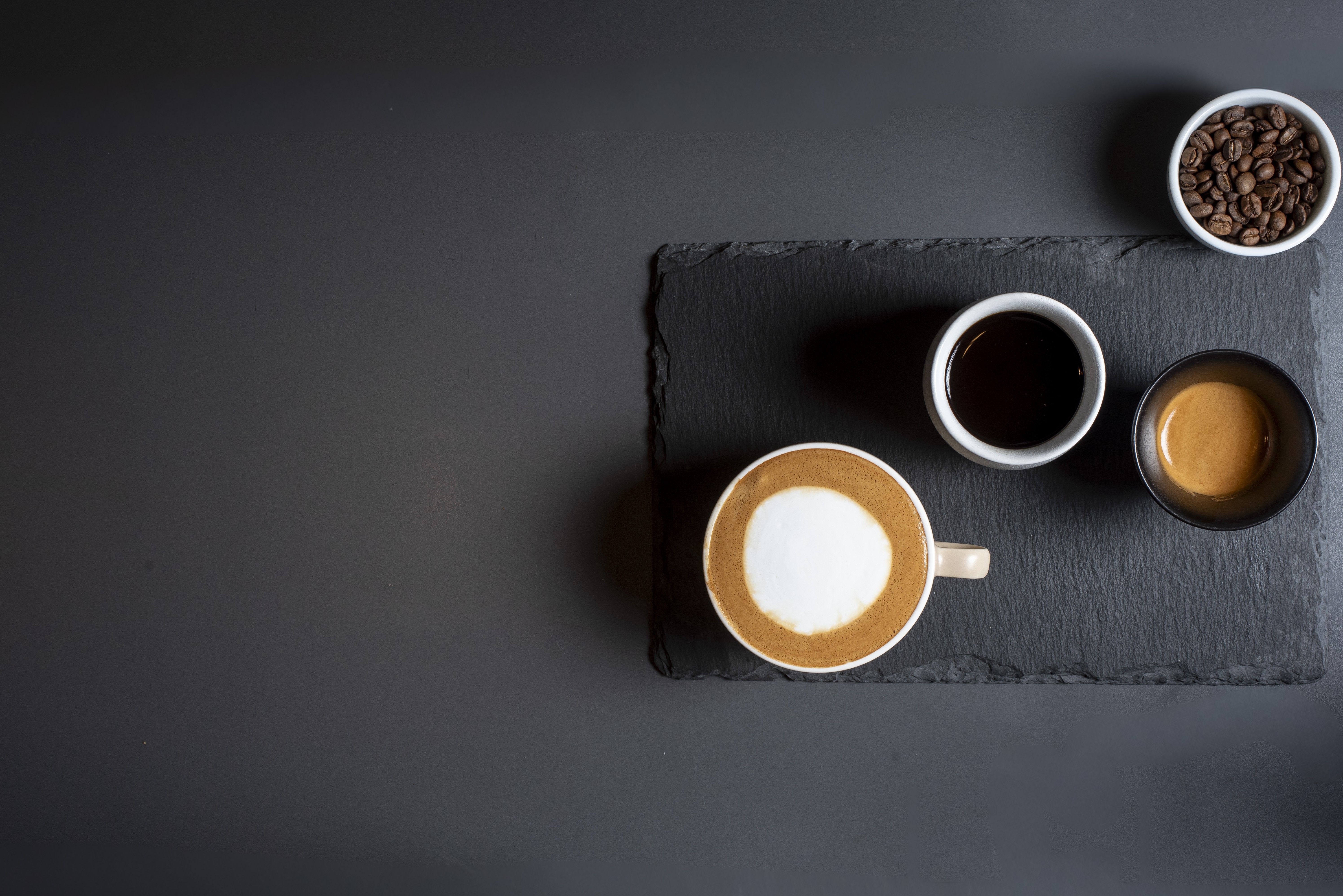 Disposi&ccedil;&atilde;o de caf&eacute;s em uma superf&iacute;cie escura: uma x&iacute;cara de cappuccino com arte de latte, uma x&iacute;cara de caf&eacute; preto, uma tigela de espresso e uma tigela com gr&atilde;os de caf&eacute; torrados, tudo sobre uma t&aacute;bua de ard&oacute;sia preta