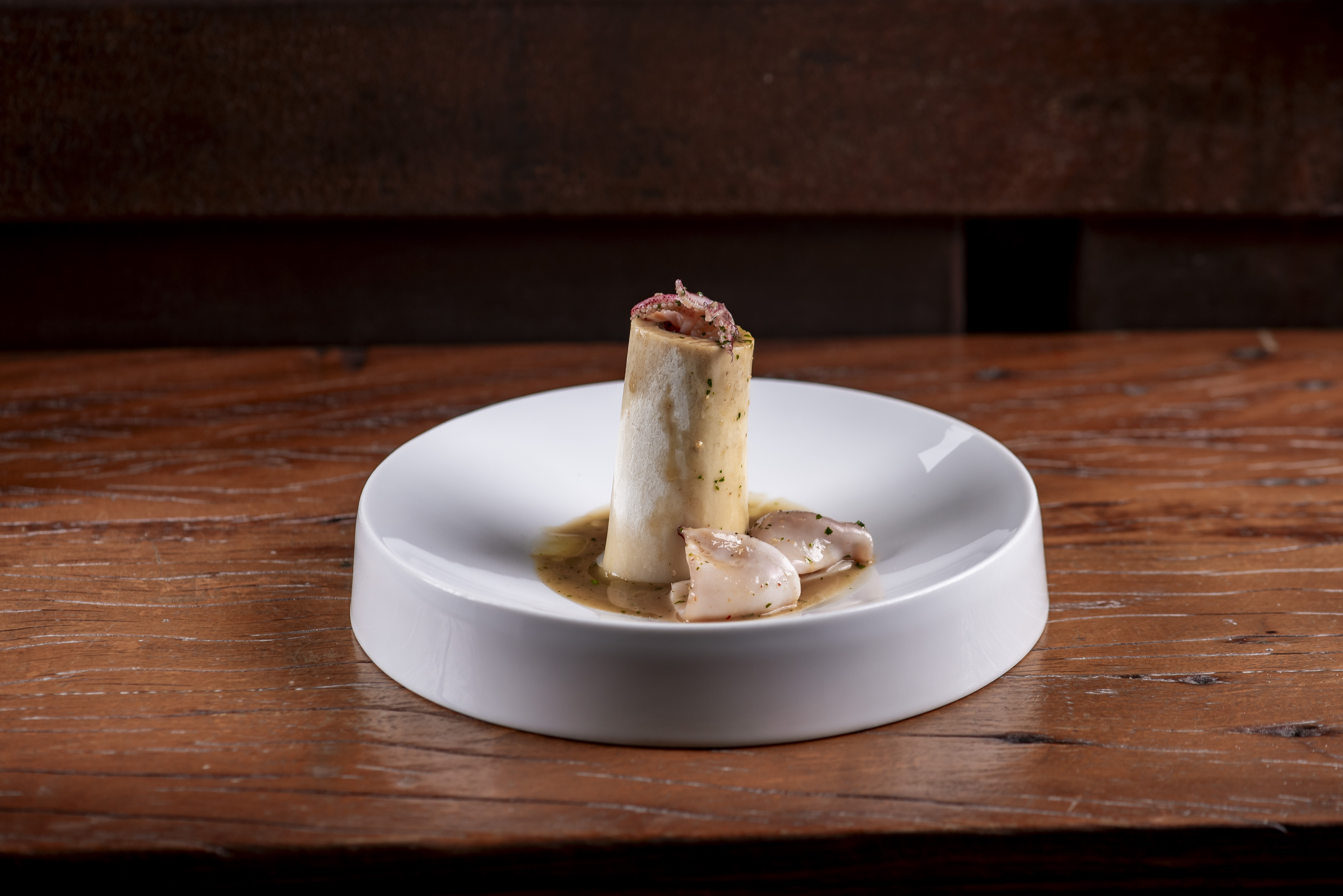 Prato gourmet com um osso cil&iacute;ndrico de tutano em p&eacute;, recheado com carne desfiada, servido em um molho cremoso com cogumelos em um prato branco sobre uma mesa de madeira r&uacute;stica