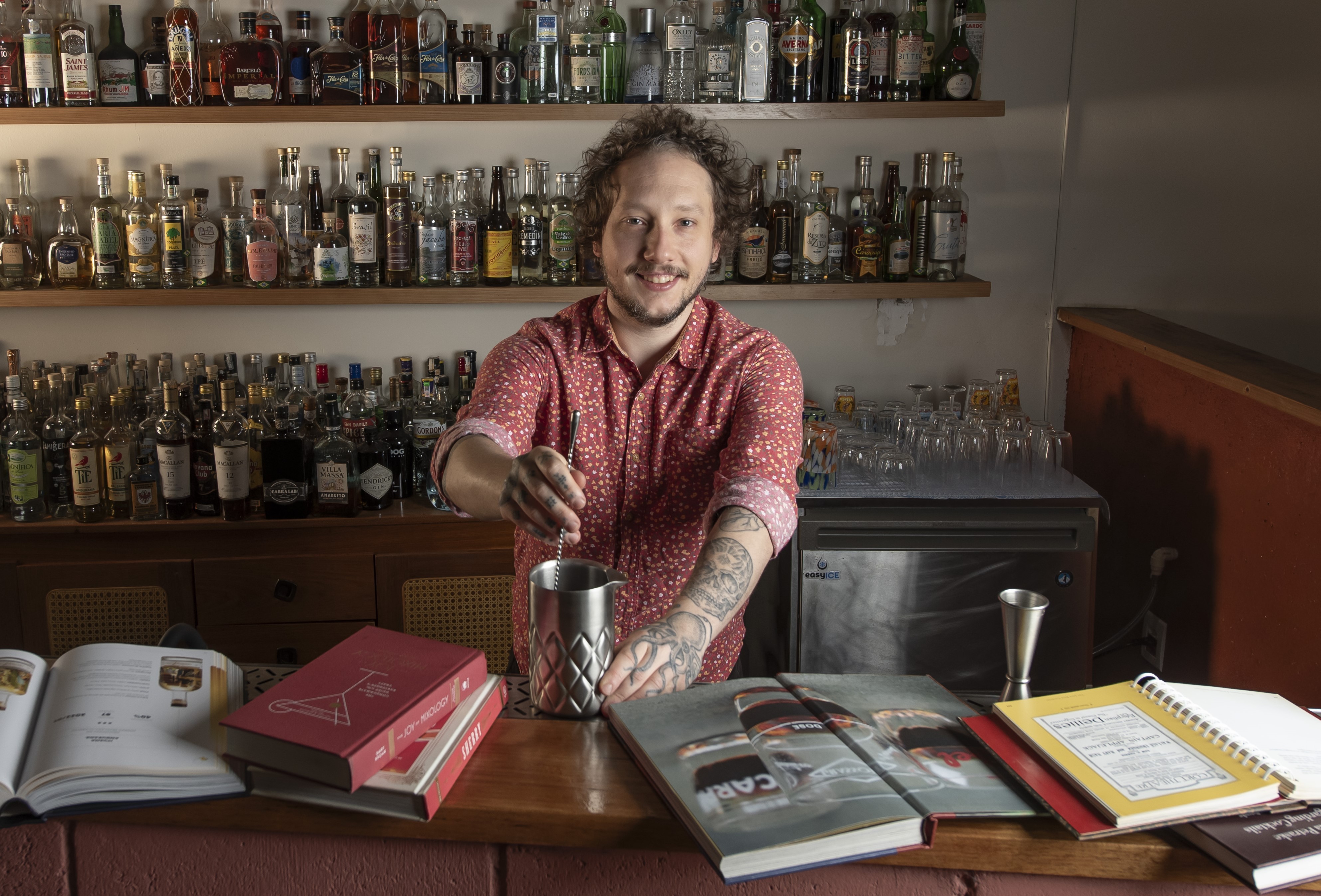 Homem branco de cabelo cacheado e barba, sorrindo, vestindo camisa vermelha estampada e com bra&ccedil;os tatuados, preparando um drink em um balc&atilde;o de bar com diversas garrafas de bebidas em prateleiras ao fundo e livros de receitas sobre o balc&atilde;o
