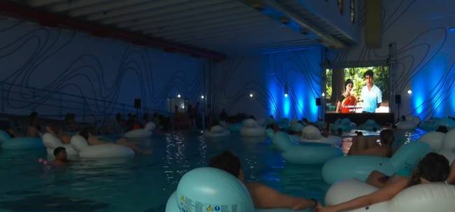 Pessoas assistem a um filme projetado em uma tela grande, flutuando em boias brancas e azuis dentro de uma piscina coberta com ilumina&ccedil;&atilde;o azul