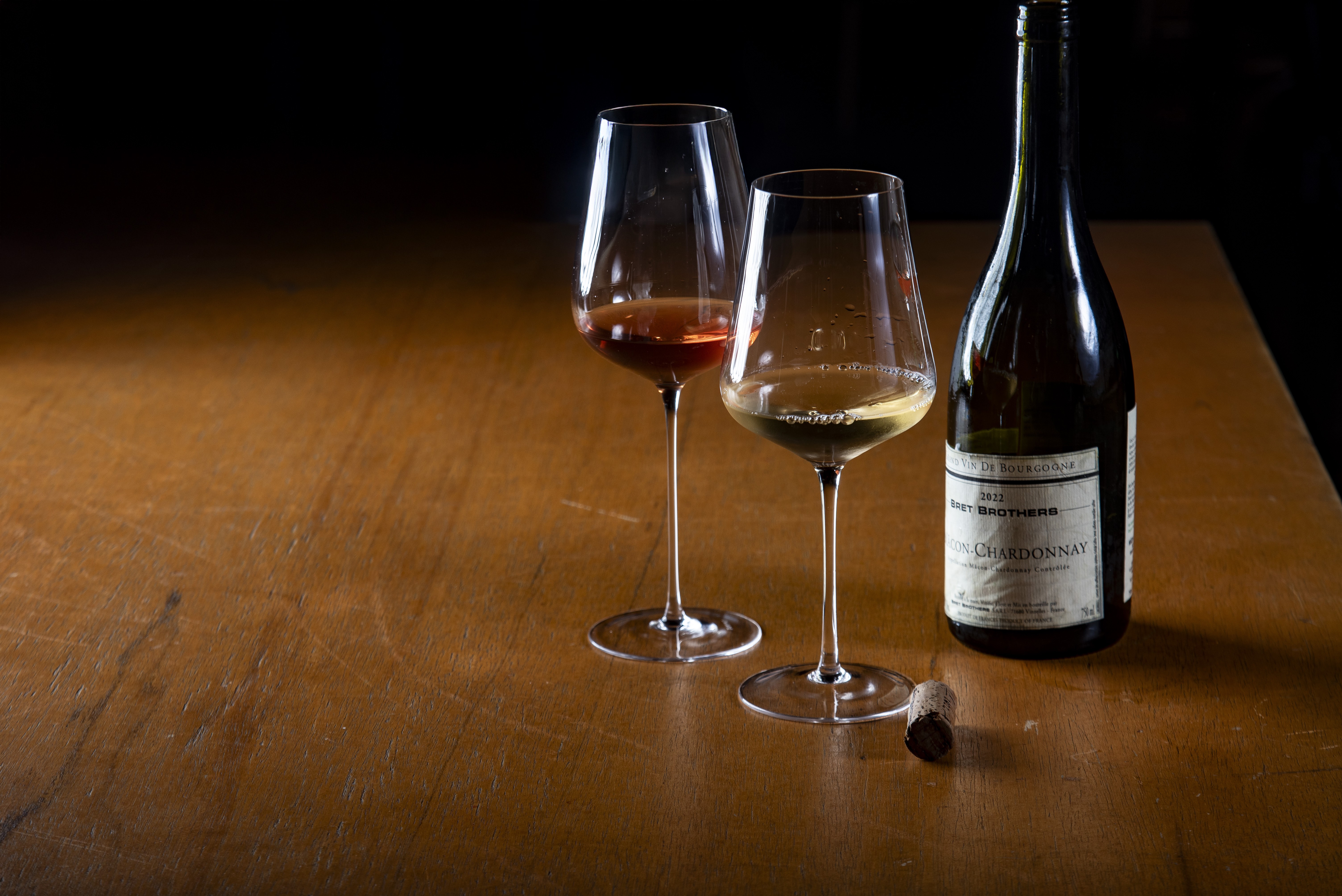 Dois c&aacute;lices de vinho, um com vinho ros&eacute; e outro com vinho branco, ao lado de uma garrafa de vinho branco e uma rolha sobre uma mesa de madeira