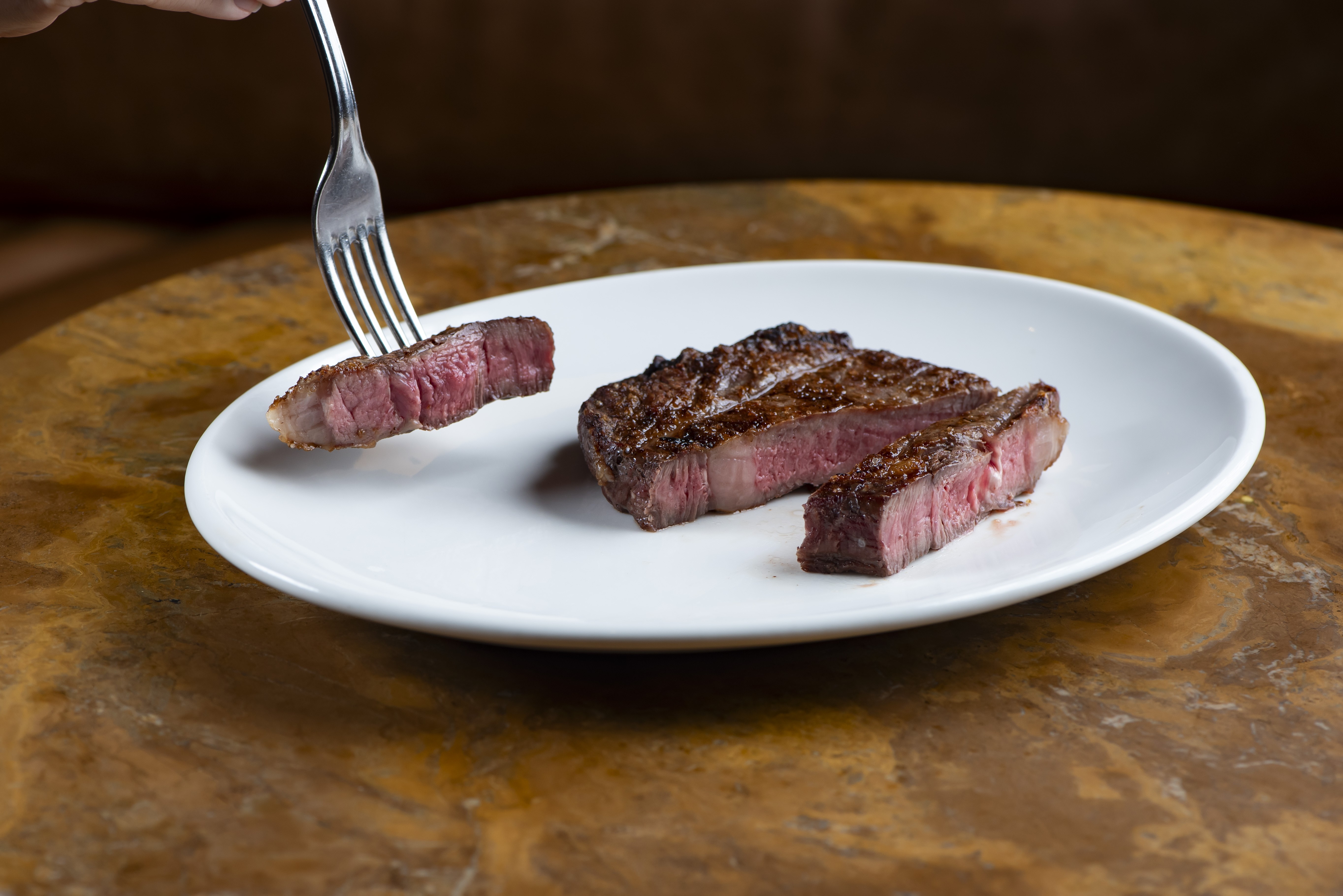 Um garfo prateado segura um peda&ccedil;o de carne vermelha malpassada sobre um prato branco. No prato, h&aacute; outro peda&ccedil;o maior de carne cortado ao meio, revelando o interior rosado. O prato est&aacute; sobre uma mesa de madeira escura com veios claros
