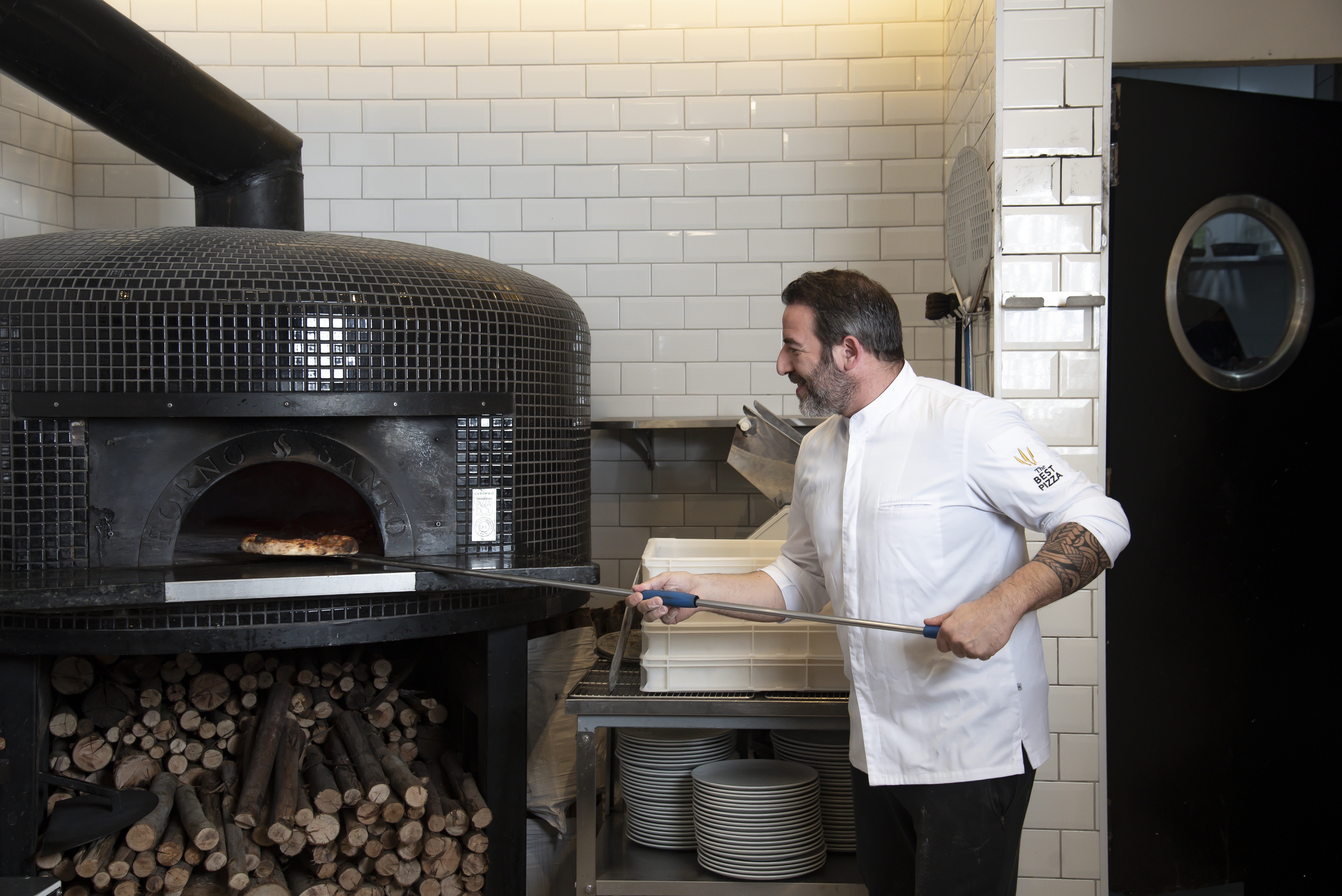 Chef sorridente com barba, vestindo d&oacute;lm&atilde; branco, usando p&aacute; met&aacute;lica para manusear uma pizza em forno a lenha preto. Lenha empilhada sob o forno, e pilhas de pratos ao lado.