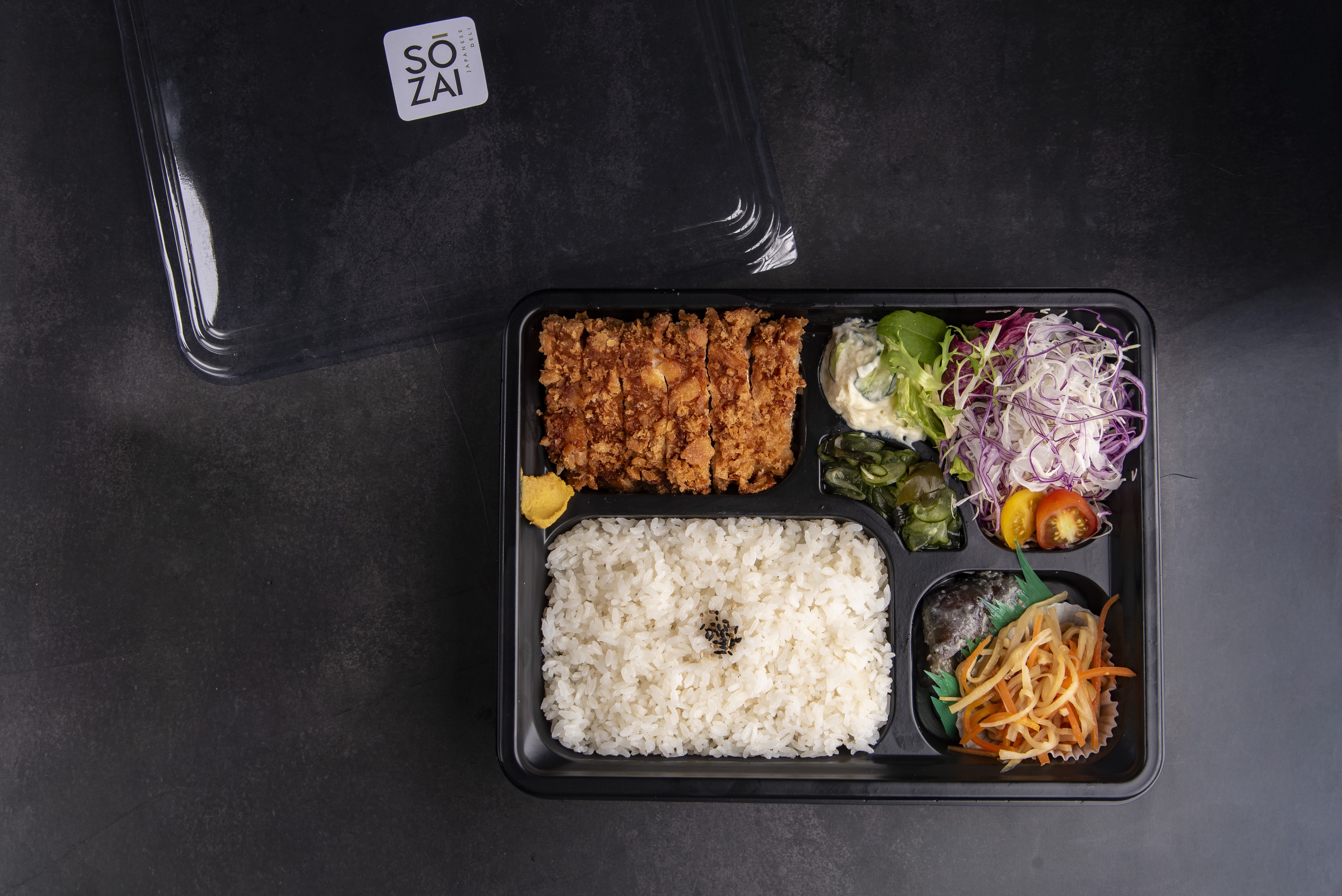 Bento box com tonkatsu fatiado, arroz branco, salada de repolho roxo e branco, salada de batata, algas e tsukemono de cenoura e nabo, sobre fundo escuro com tampa de pl&aacute;stico transparente e adesivo S&Ocirc; ZAI