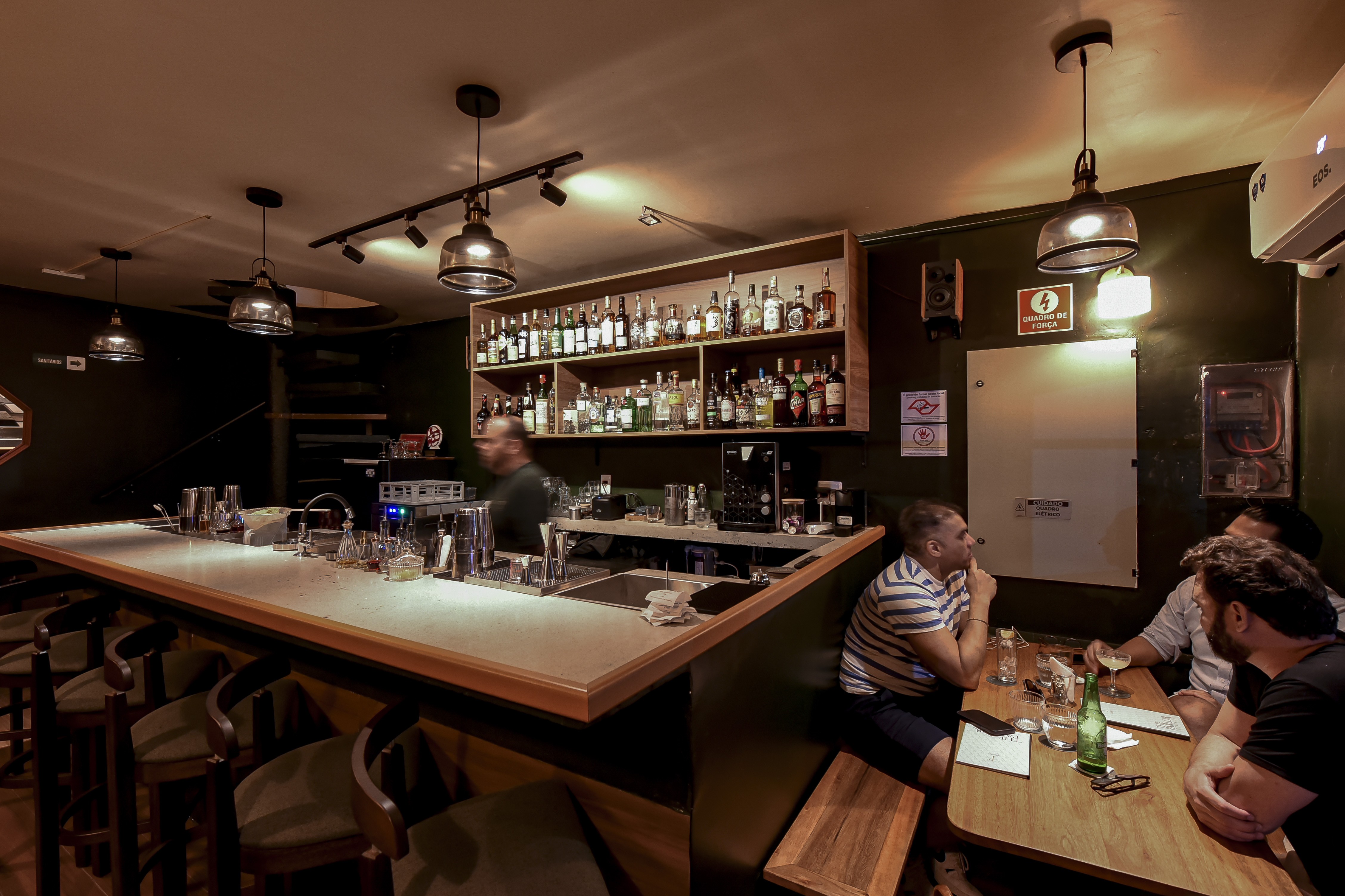 Interior de um bar com ilumina&ccedil;&atilde;o baixa, balc&atilde;o comprido &agrave; esquerda com bancos, e prateleiras de bebidas ao fundo. Um bartender trabalha no balc&atilde;o, e &agrave; direita, tr&ecirc;s homens conversam sentados em uma mesa de madeira, com bebidas e copos.