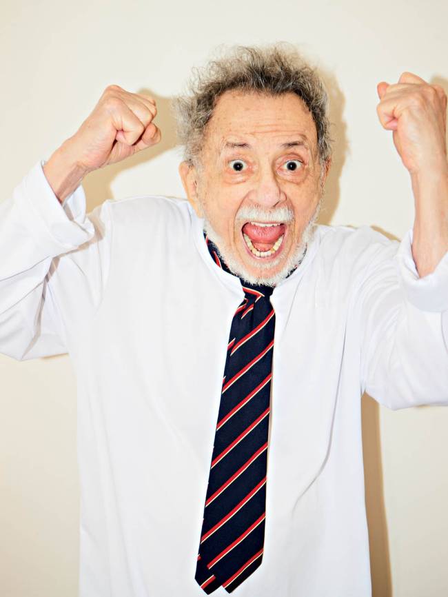 Homem idoso de cabelos grisalhos e barba branca, vestindo camisa social branca e gravata azul-marinho com listras vermelhas e brancas, comemora com os punhos cerrados para cima e a boca aberta em um grito de alegria, em fundo claro