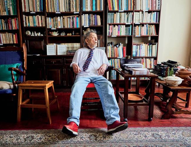 Homem idoso de cabelos grisalhos e camisa branca com gravata listrada azul e vermelha, cal&ccedil;a jeans e t&ecirc;nis vermelhos, sentado em uma poltrona de madeira. Ele olha para a direita, em um ambiente com estantes cheias de livros e m&oacute;veis de madeira com objetos antigos
