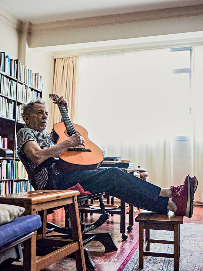 Homem idoso de barba grisalha, camiseta listrada e cal&ccedil;a escura, sentado em uma cadeira, tocando viol&atilde;o. Ele tem os p&eacute;s cal&ccedil;ados com t&ecirc;nis vermelhos apoiados em um banquinho de madeira. Ao fundo, uma estante cheia de livros e uma janela com cortinas claras. O ambiente &eacute; iluminado e aconchegante