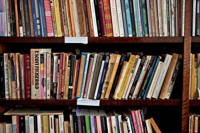 Estantes de madeira escura repletas de livros antigos e novos, com lombadas de diversas cores e tamanhos, alguns inclinados, em uma biblioteca ou livraria