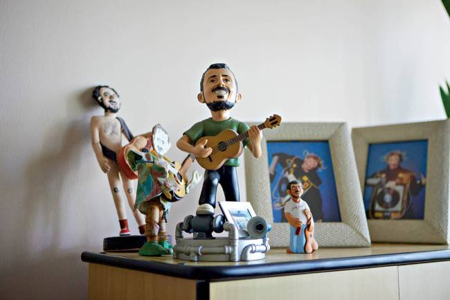 Uma cole&ccedil;&atilde;o de bonecos e porta-retratos sobre uma mesa de madeira. No centro, um boneco de um homem barbudo tocando viol&atilde;o. &Agrave; esquerda, um boneco nu e outro colorido. &Agrave; direita, dois porta-retratos com fotos de pessoas e um boneco pequeno. O fundo &eacute; uma parede branca
