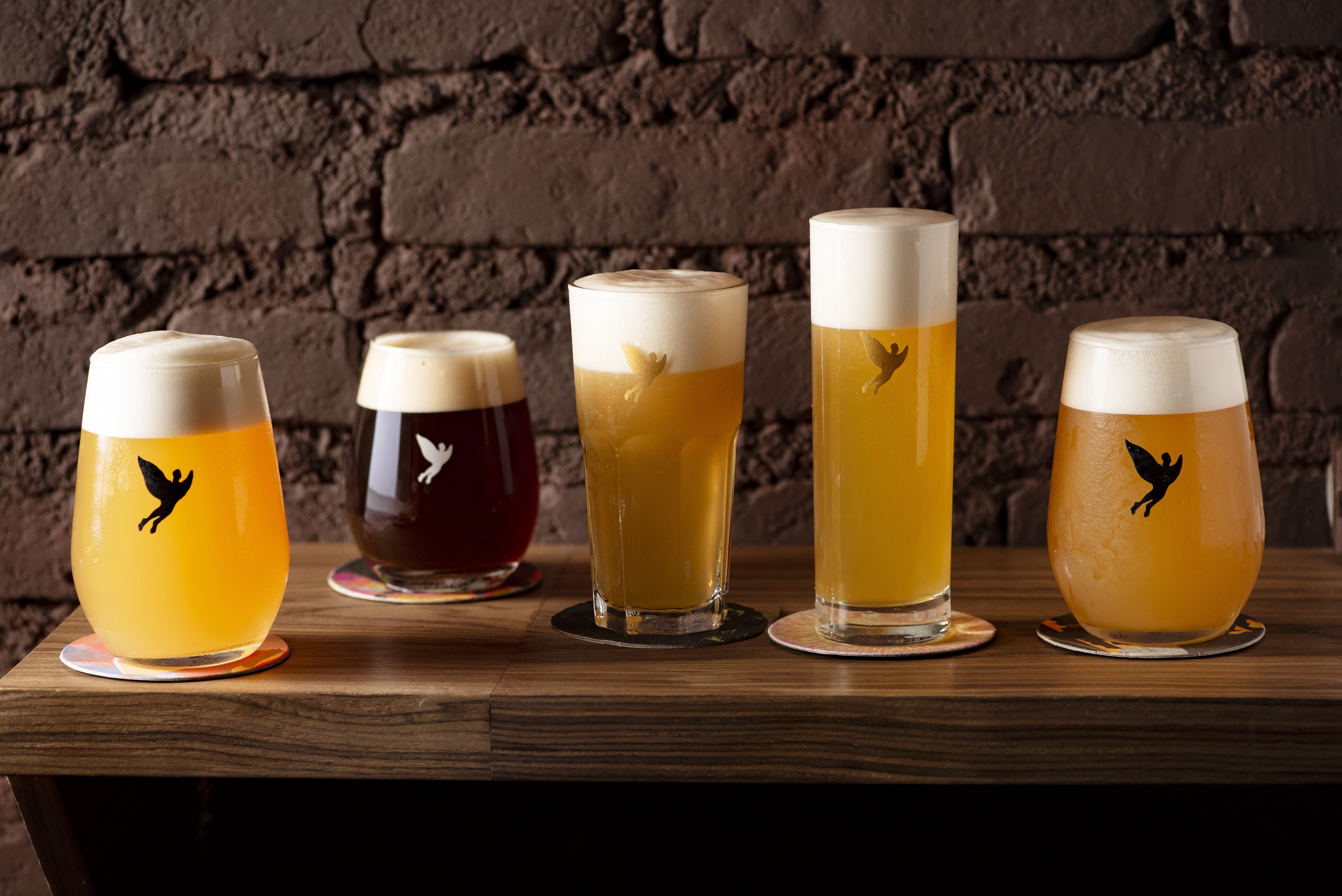 Cinco copos de cerveja artesanal com espuma branca, tr&ecirc;s claras e duas escuras, sobre uma bancada de madeira r&uacute;stica, com um fundo de parede de tijolos escuros. Cada copo tem um adesivo de p&aacute;ssaro voando