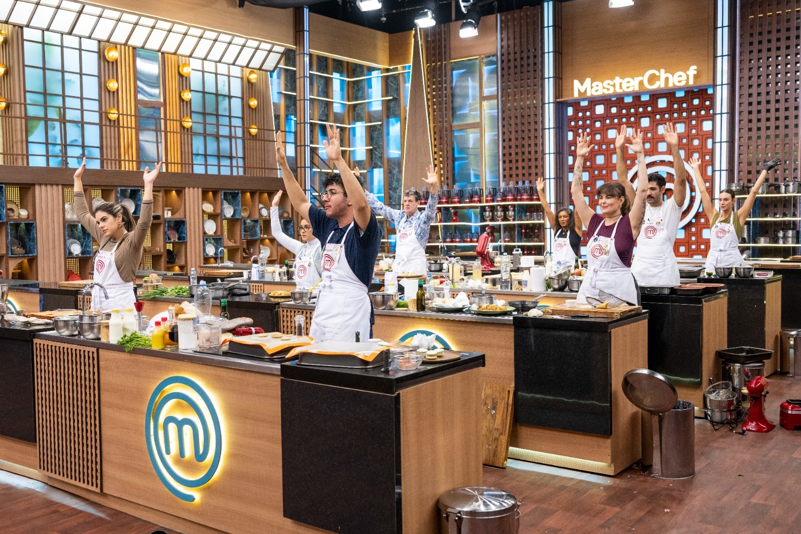 Competidores do MasterChef, usando aventais brancos, levantam os bra&ccedil;os em uma cozinha profissional. Homens e mulheres est&atilde;o em bancadas individuais com ingredientes e utens&iacute;lios, sob o letreiro MasterChef ao fundo. O ambiente &eacute; iluminado, com detalhes em madeira e metal.