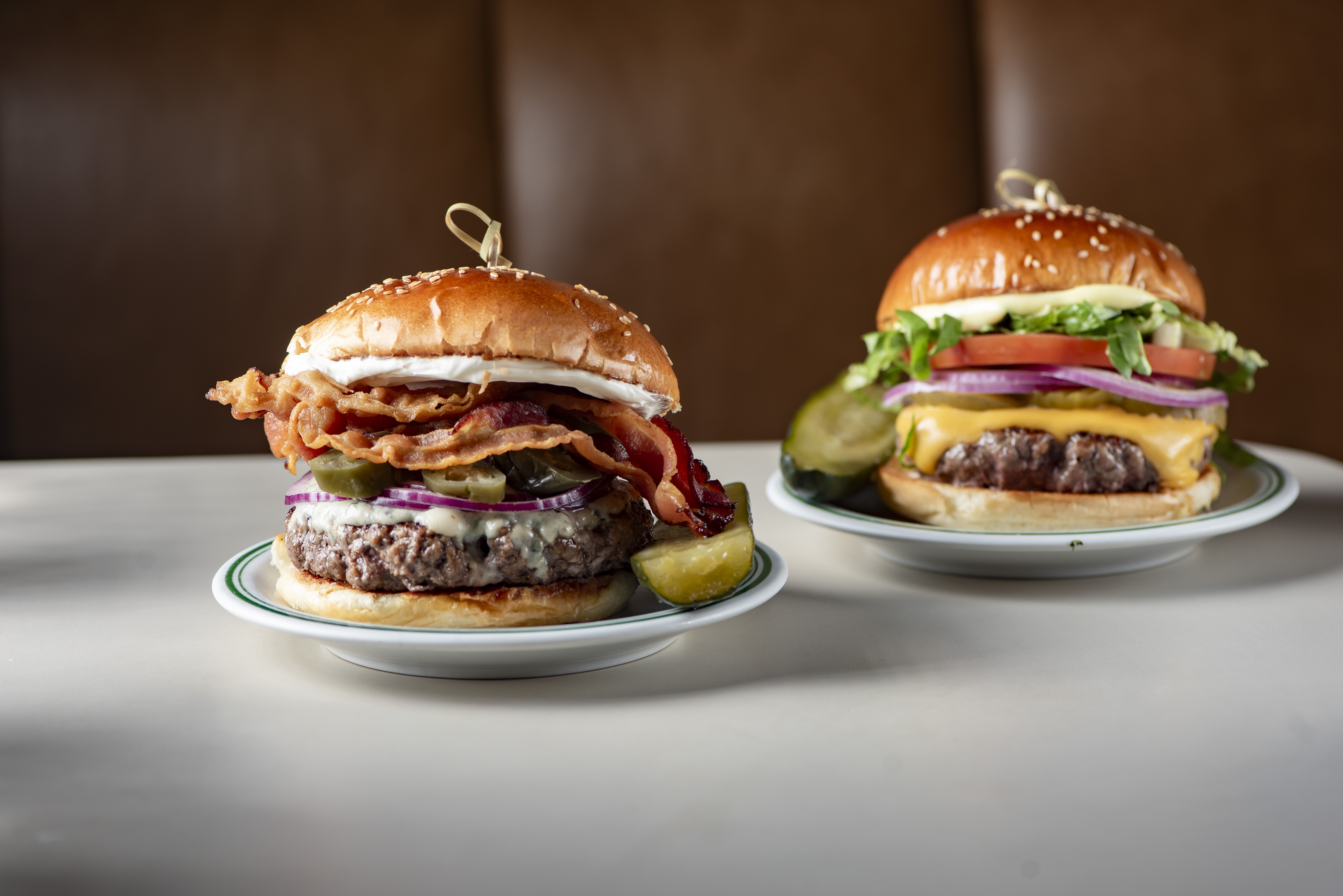 Dois hamb&uacute;rgueres em pratos brancos com borda verde sobre uma mesa clara. O da esquerda tem bacon crocante, queijo azul, cebola roxa, picles e um gomo de lim&atilde;o. O da direita tem queijo cheddar derretido, alface, tomate, cebola roxa e picles
