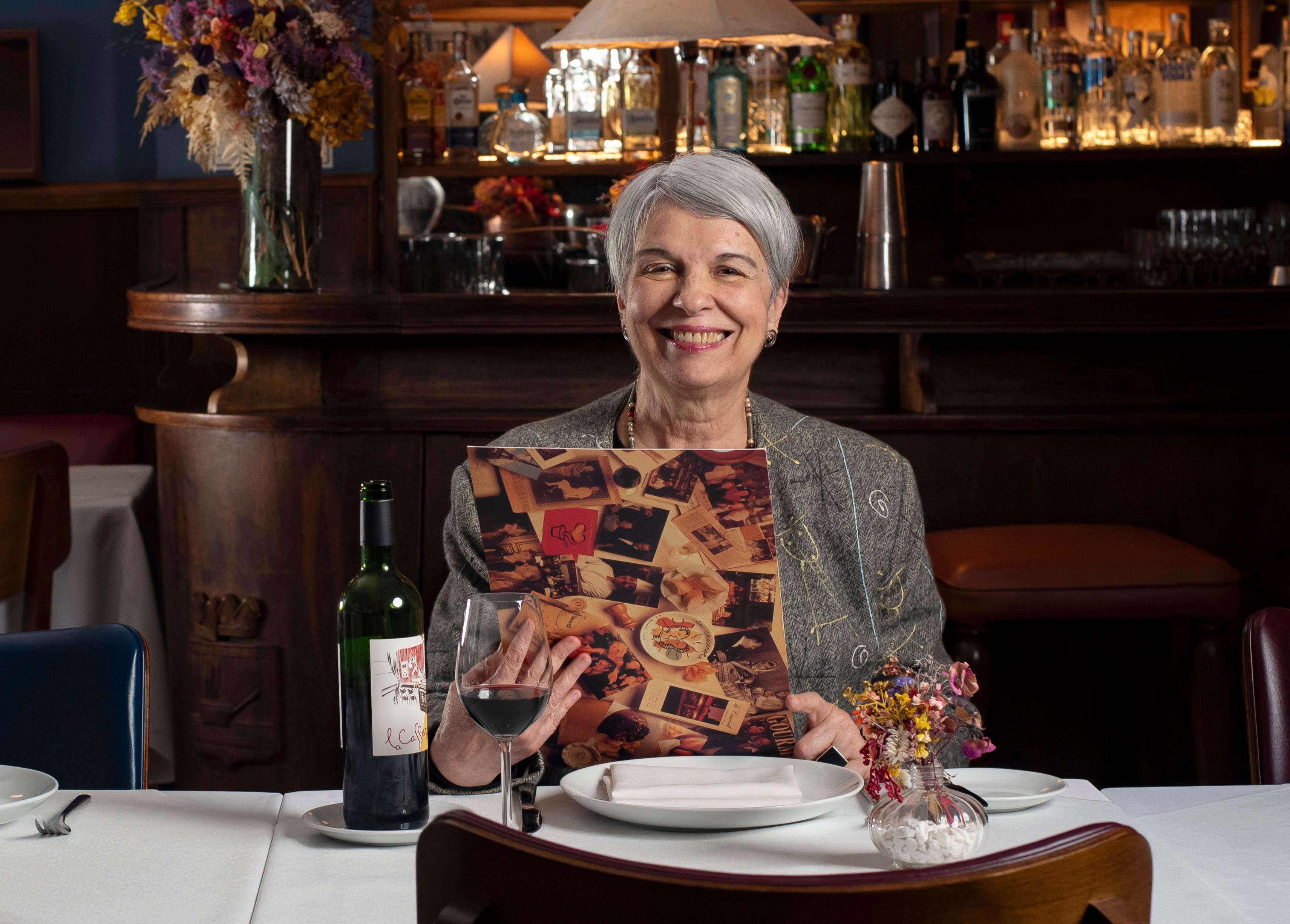 Mulher de cabelos grisalhos e blazer estampado sorri, segurando um card&aacute;pio com fotos de pratos, sentada &agrave; mesa de um restaurante com uma garrafa e ta&ccedil;a de vinho, e um arranjo floral. Ao fundo, um bar com garrafas de bebidas