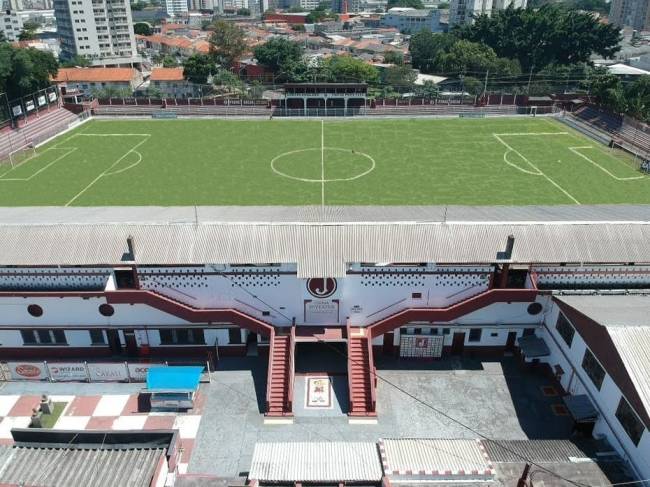 Vista a&eacute;rea de um est&aacute;dio de futebol com gramado verde e marca&ccedil;&otilde;es brancas. Abaixo, um edif&iacute;cio branco e vermelho com escadarias sim&eacute;tricas que levam a um p&aacute;tio. Ao fundo, pr&eacute;dios e casas da cidade sob c&eacute;u claro