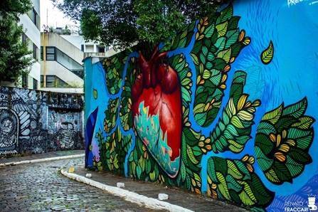 Mural vibrante em rua de paralelep&iacute;pedos, com um cora&ccedil;&atilde;o vermelho e azul-claro brotando folhas verdes em um fundo azul-escuro. Ao lado, outro muro com grafites em preto e branco, e edif&iacute;cios ao fundo